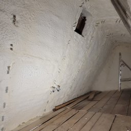 Tatrapiana Daniel Czyszczoń - Poddasze po ociepleniu pianką PUR, widoczne drewniane deski podłogowe, fragment okna i drabina. Biała, nierówna powierzchnia pianki pokrywa skośne ściany.