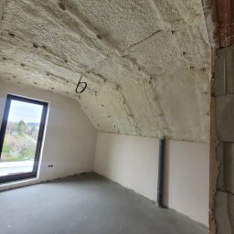 Tatrapiana Daniel Czyszczoń - Poddasze po natrysku izolacji pianką PUR: widoczne ocieplenie na skosach dachu, okno z widokiem na zewnątrz, surowy beton na podłodze i fragment ściany z pustaków.