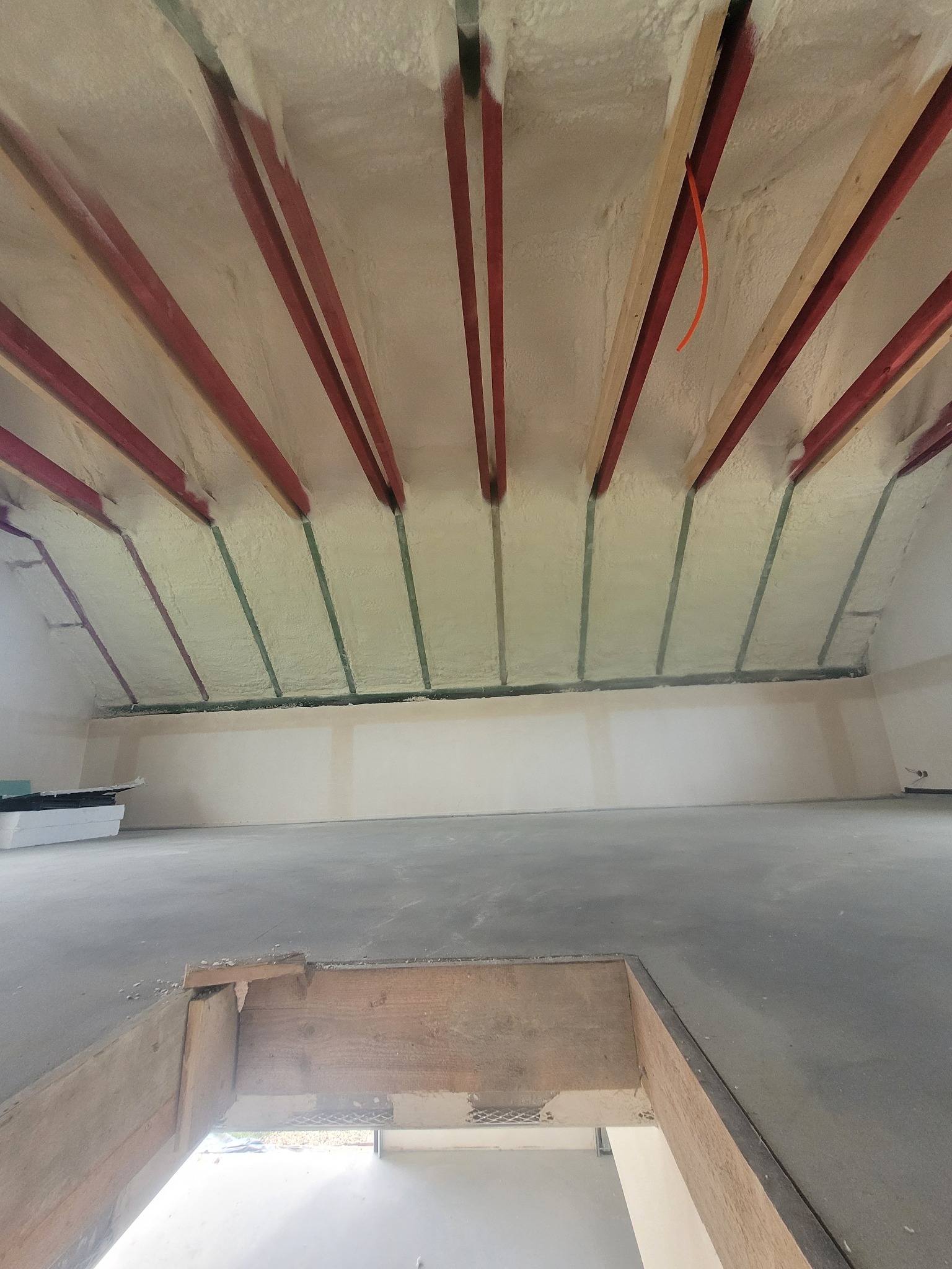 Ocieplone poddasze z widocznymi elementami konstrukcyjnymi. Izolacja natryskowa między krokwiami. Surowy beton na podłodze. Widok z dołu na ocieplony strop.