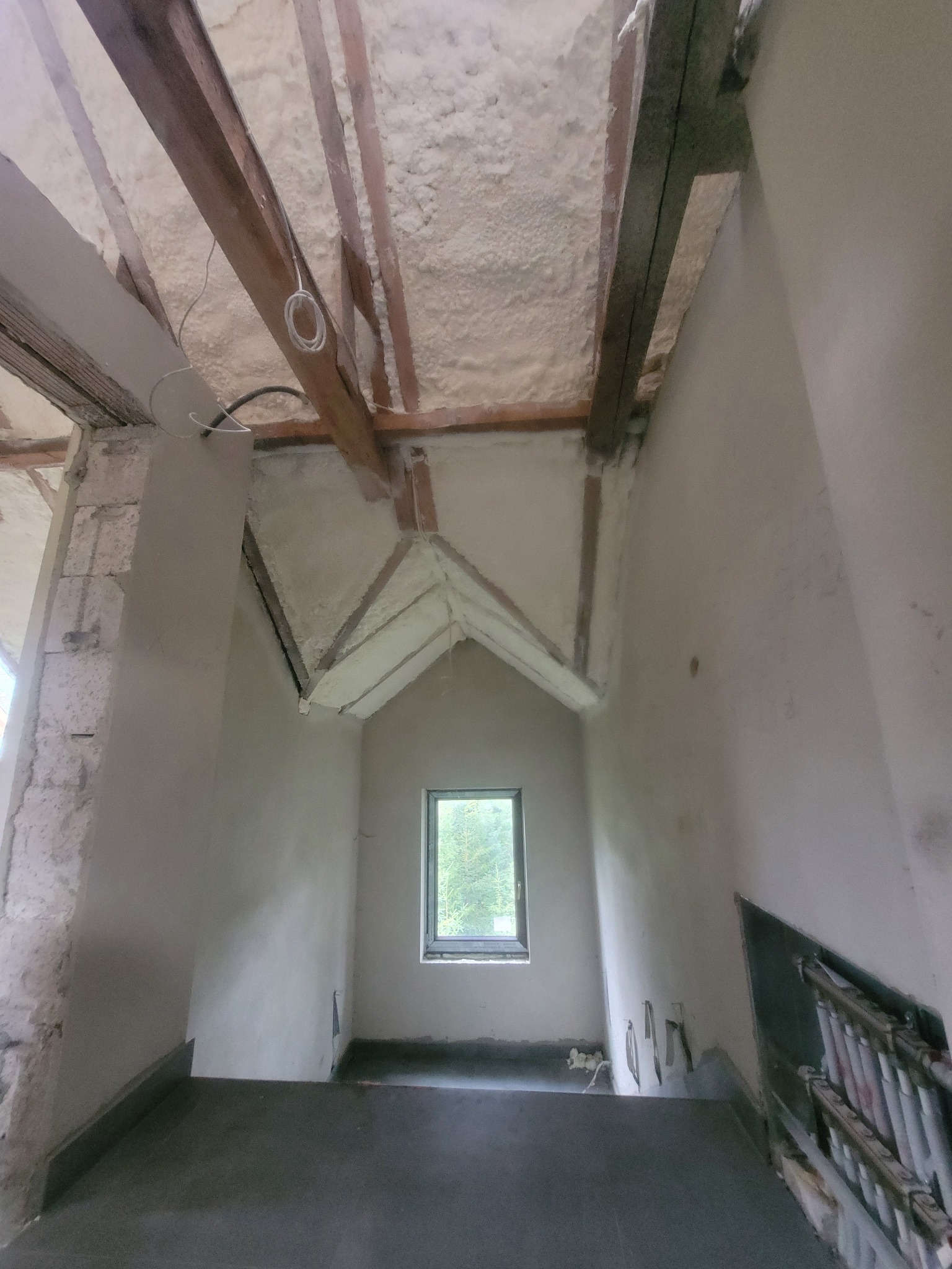 Poddasze z oknem po natrysku pianki PUR, widoczne drewniane elementy konstrukcyjne i ocieplone skosy. Świeża warstwa izolacji.