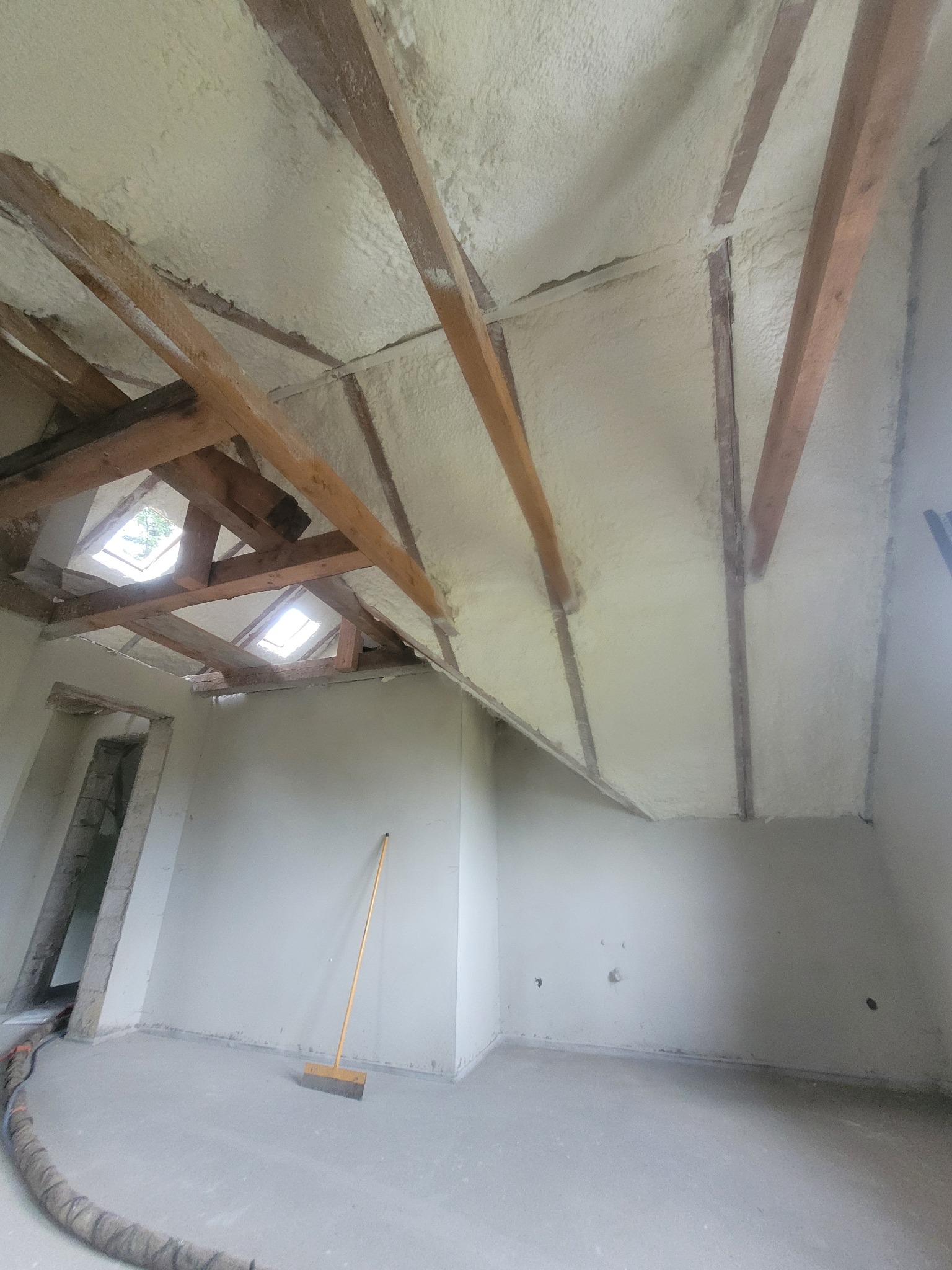 Ocieplone poddasze pianką PUR z widocznymi drewnianymi belkami. W pomieszczeniu stoi miotła, a przez okna dachowe wpada światło.