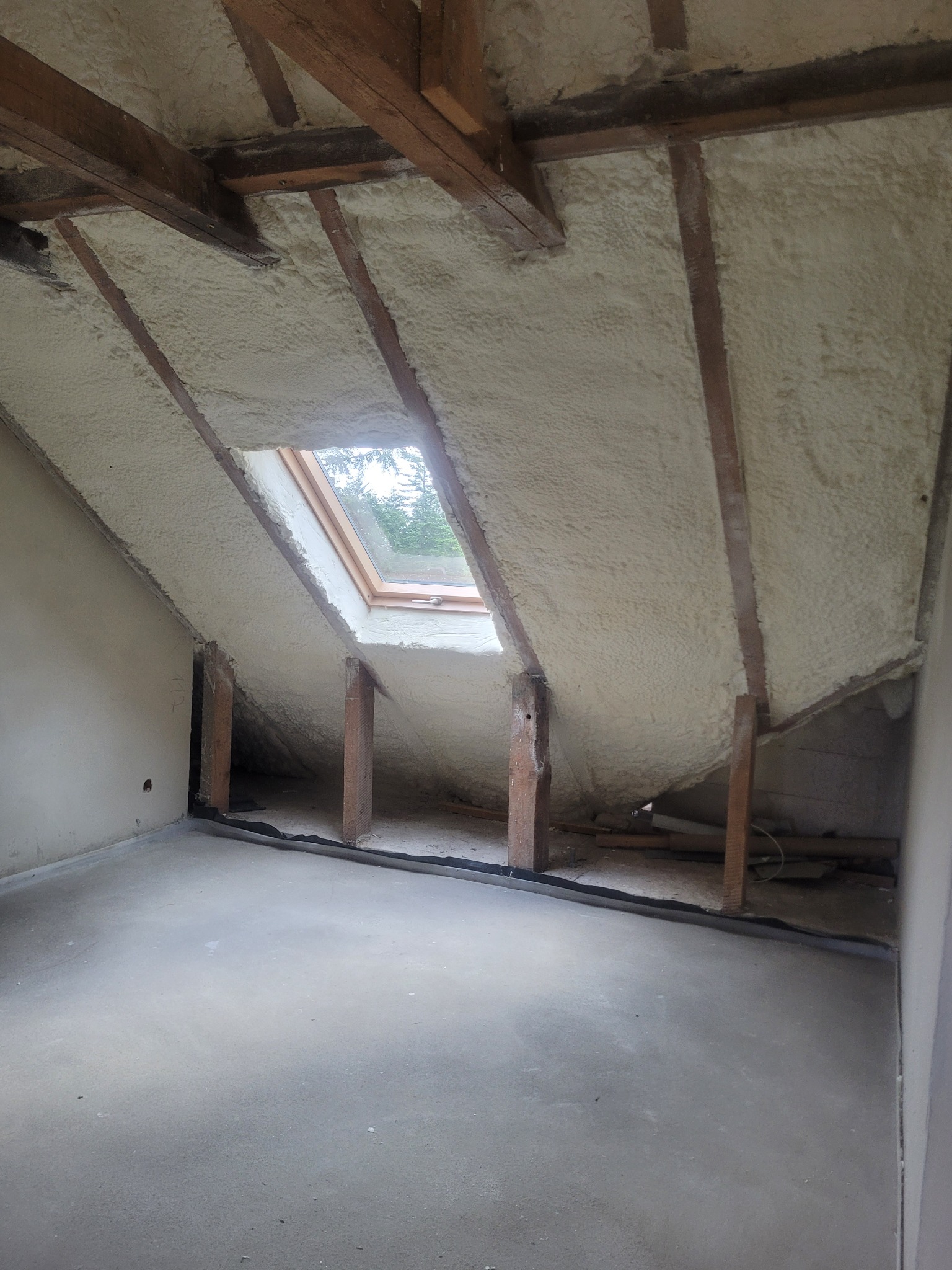 Poddasze z oknem połaciowym po natrysku izolacji pianką. Widoczne drewniane elementy konstrukcyjne i równomierna warstwa ocieplenia. Surowy beton na podłodze.