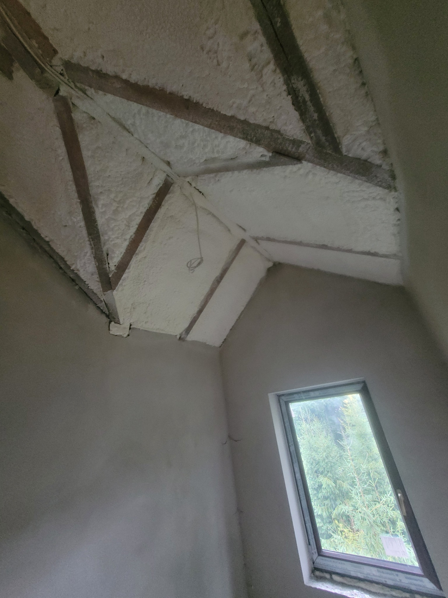 Poddasze po ociepleniu pianką PUR: widoczne drewniane elementy konstrukcyjne i biała pianka. W tle okno z widokiem na drzewa. Ściany w stanie surowym.