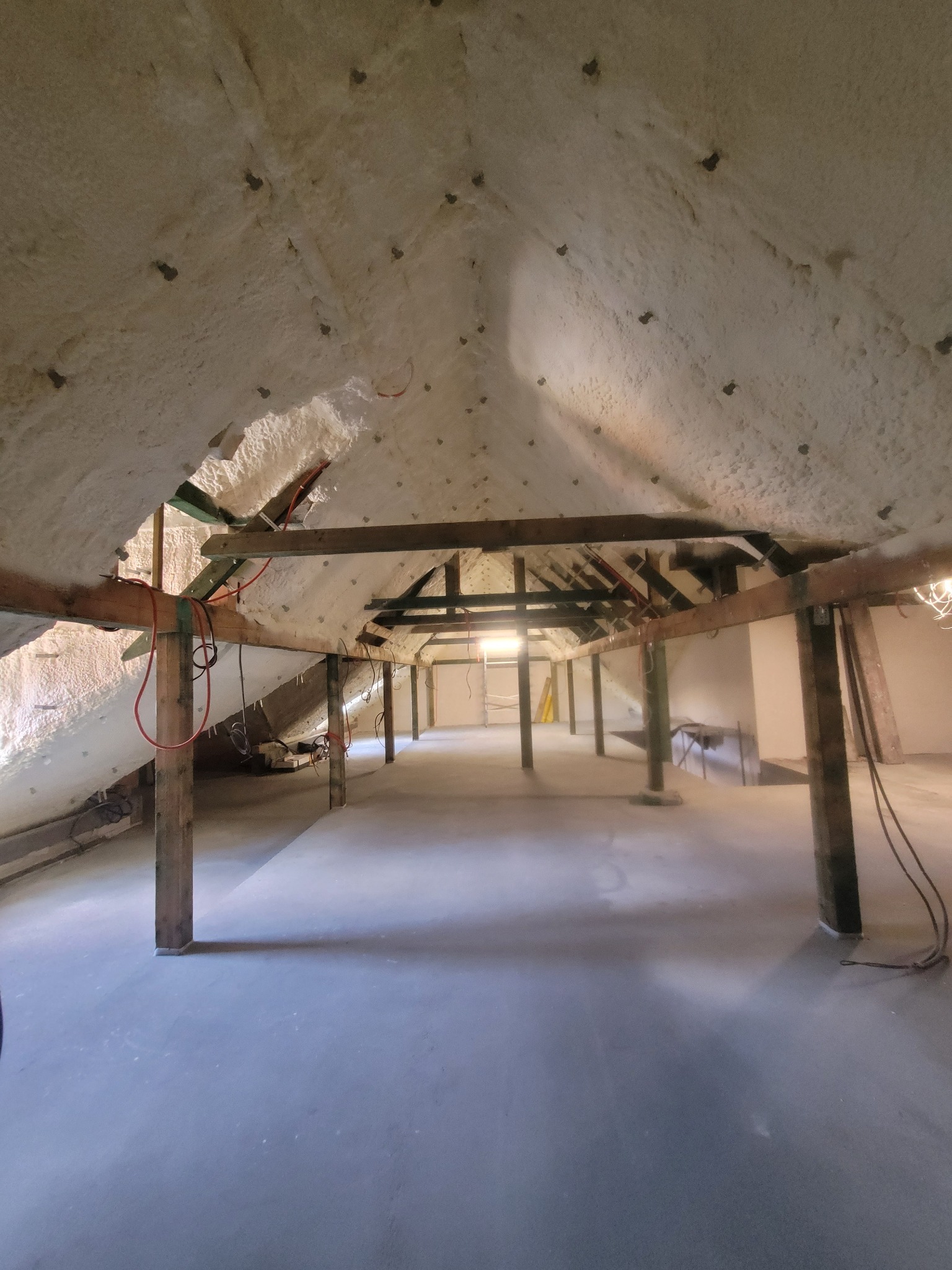 Poddasze po natrysku izolacji piankowej, widoczne drewniane elementy konstrukcyjne i równomierna warstwa piany na skośnych powierzchniach dachu. Widok z dołu, surowy stan.