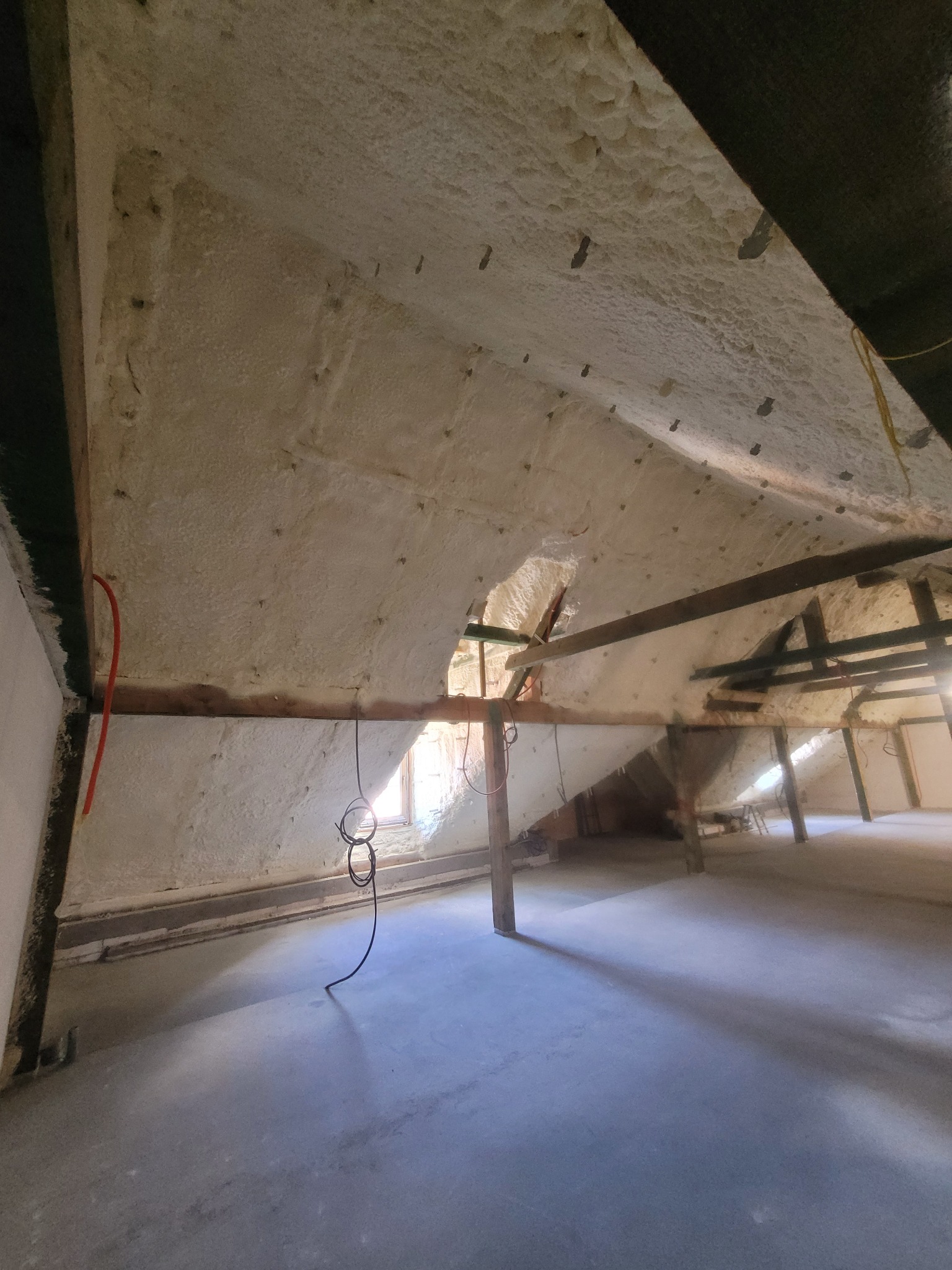 Poddasze po natrysku izolacji. Widoczne drewniane elementy konstrukcyjne i równomiernie nałożona warstwa izolacji. Światło dzienne wpada przez okno.