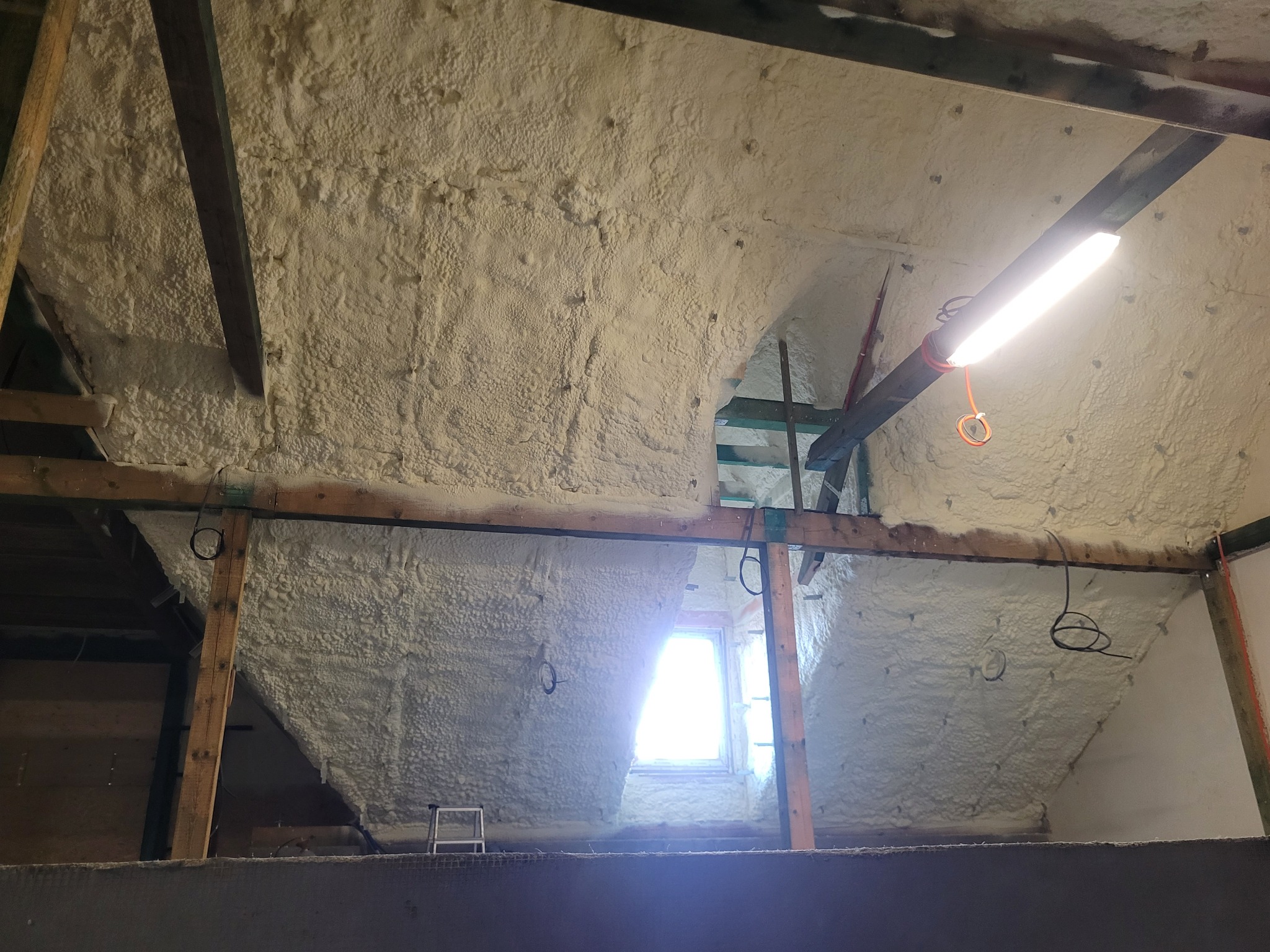 Poddasze po ociepleniu pianką PUR, widoczne drewniane elementy konstrukcyjne i okno. Oświetlenie robocze. Tekstura pianki równomierna, pokrywająca całą powierzchnię.
