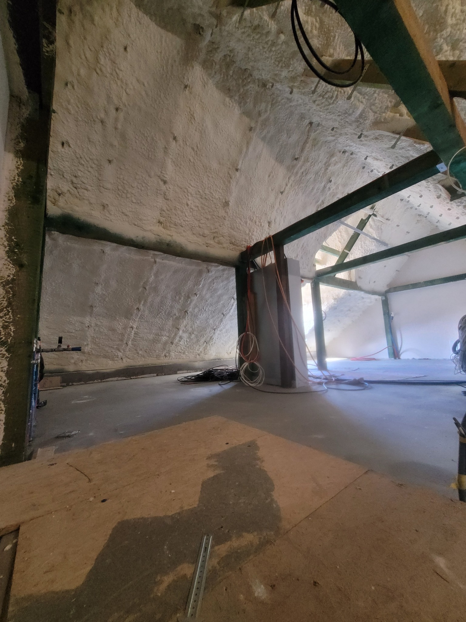 Poddasze po natrysku izolacji. Widoczne drewniane elementy konstrukcyjne i ściany pokryte grubą warstwą jasnej piany izolacyjnej. Na podłodze widoczne przewody i płyty OSB.