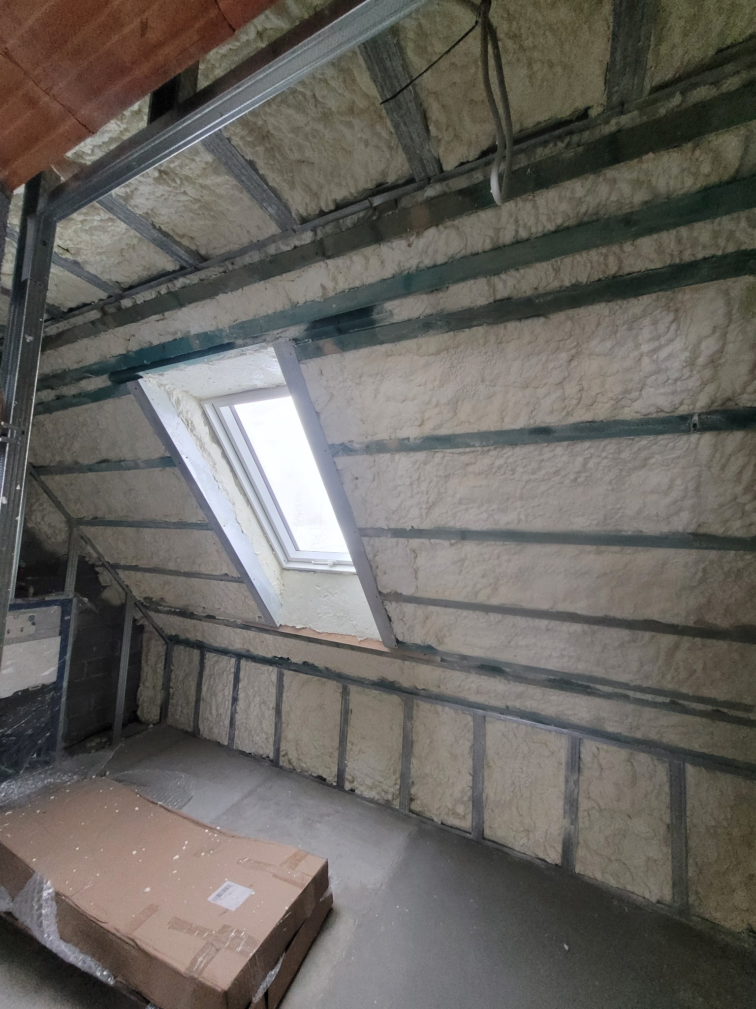 Poddasze po natrysku izolacji, widoczne metalowe profile i okno dachowe. Izolacja wypełnia przestrzeń między profilami, tworząc jednolitą warstwę. Na podłodze karton.