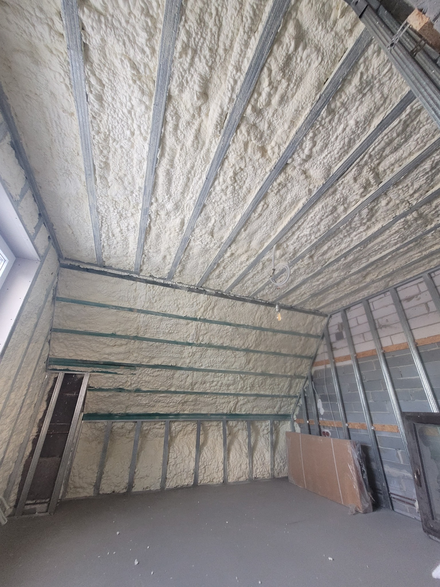 Poddasze po natrysku izolacji. Widoczne metalowe profile konstrukcyjne i równomierna warstwa piany na skośnych ścianach i suficie. Izolacja w odcieniach bieli i kremu.