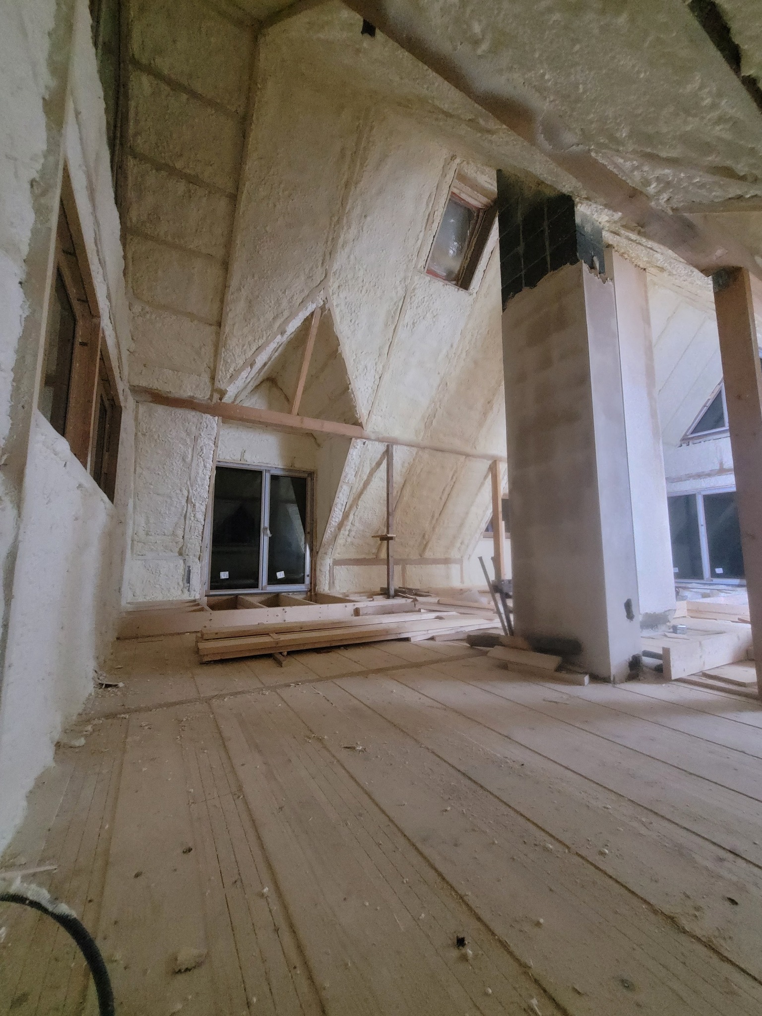 Poddasze po natrysku izolacji piankowej, widoczne okna i drewniane elementy konstrukcyjne. Ściany i sufit pokryte grubą warstwą piany. Widok z poziomu podłogi.