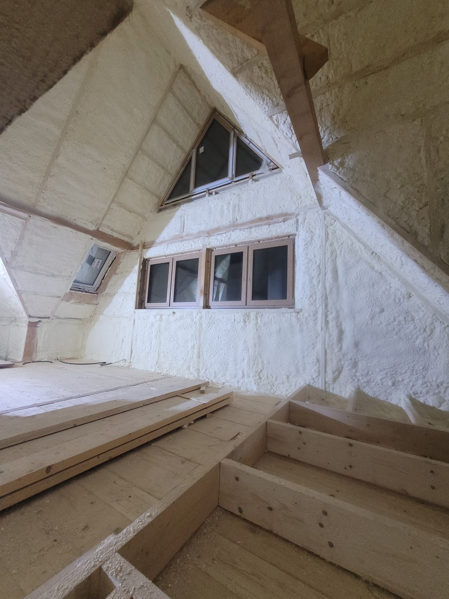 Poddasze z oknami po natrysku izolacji, widoczne drewniane elementy konstrukcyjne i podłoga. Biała, nierówna powierzchnia izolacji pokrywa ściany i sufit.