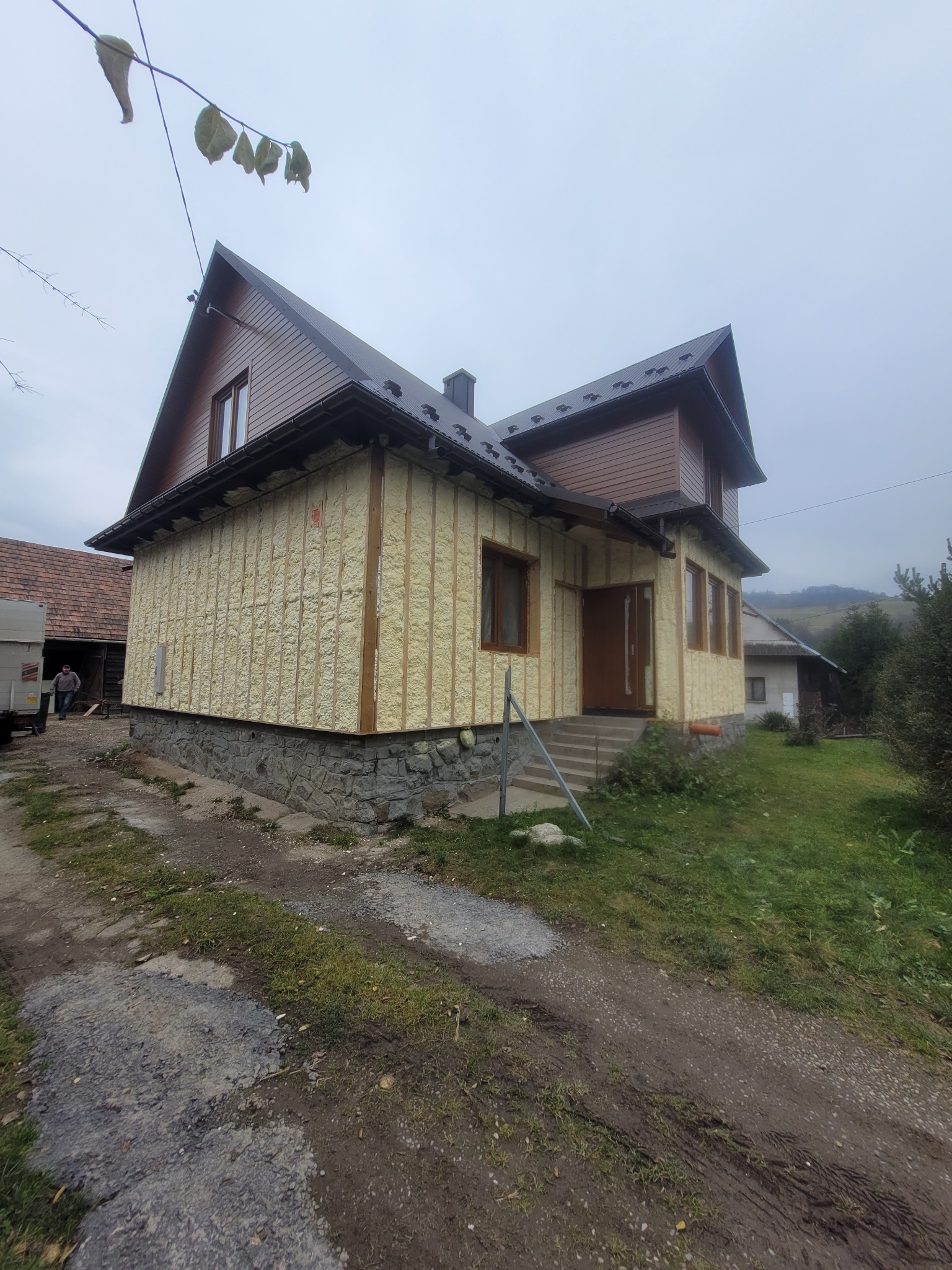 Dom z ocieploną ścianą zewnętrzną, widoczne drewniane ramy i wypełnienie pianką. Ujęcie z dołu, na pierwszym planie ścieżka i trawa. W tle zalesione wzgórza.