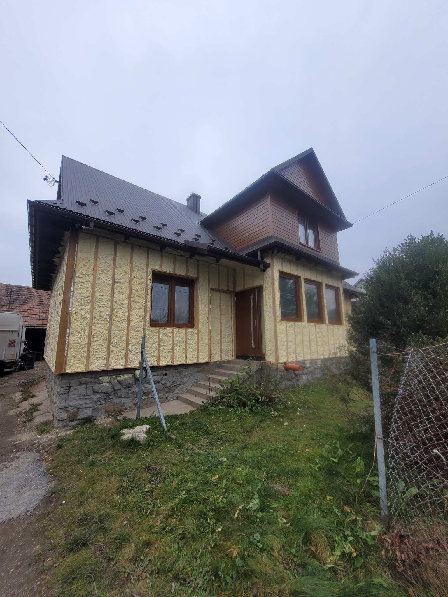 Dom w trakcie ocieplania: widoczne drewniane listwy i wypełniająca je warstwa izolacyjna. Część elewacji wykończona drewnem. Szara, pochmurna pogoda.