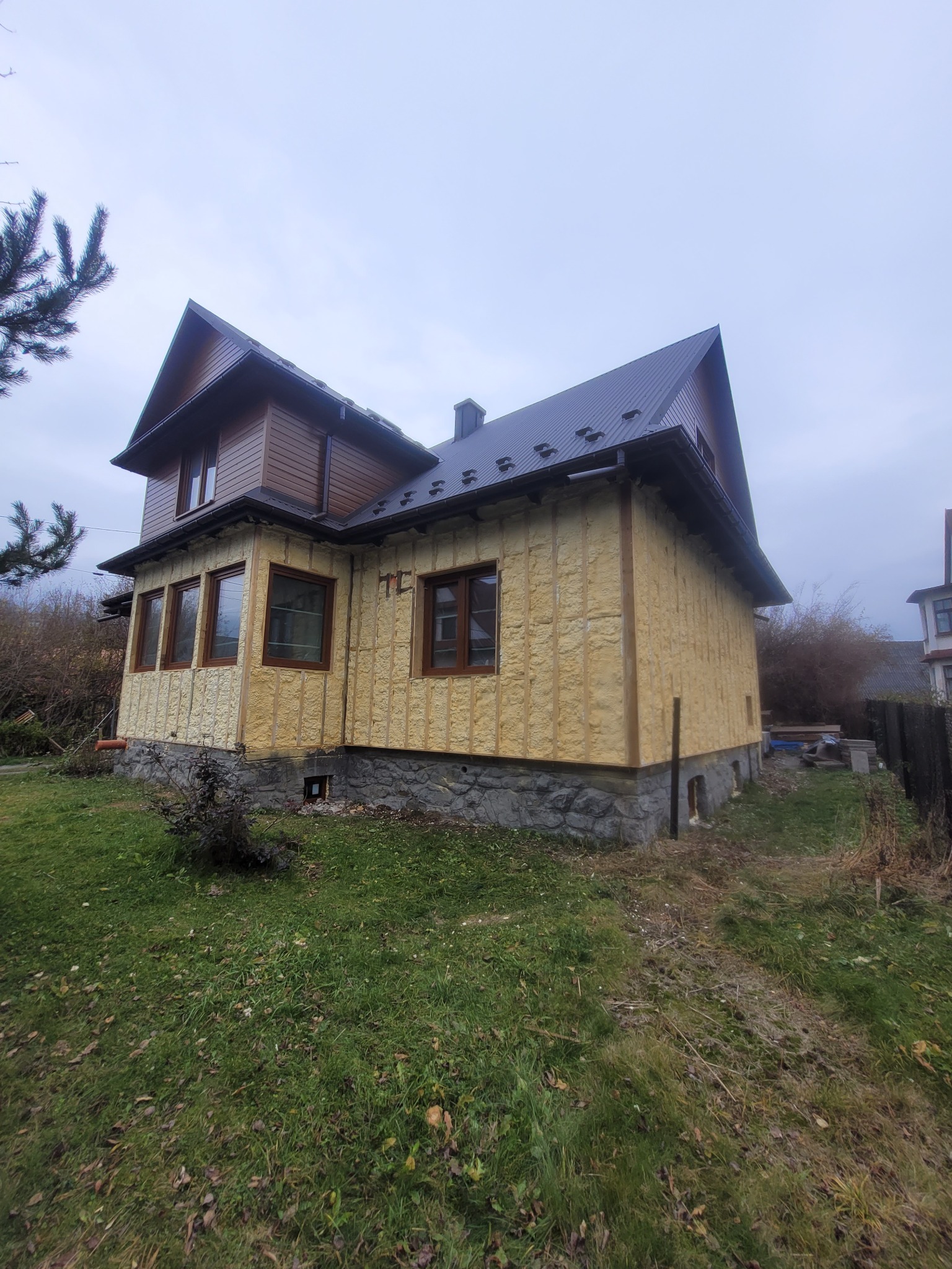 Dom jednorodzinny w trakcie ocieplania pianką PUR, widok z boku na elewację z widoczną strukturą piany i drewnianymi ramami okiennymi. Częściowo ocieplony budynek.