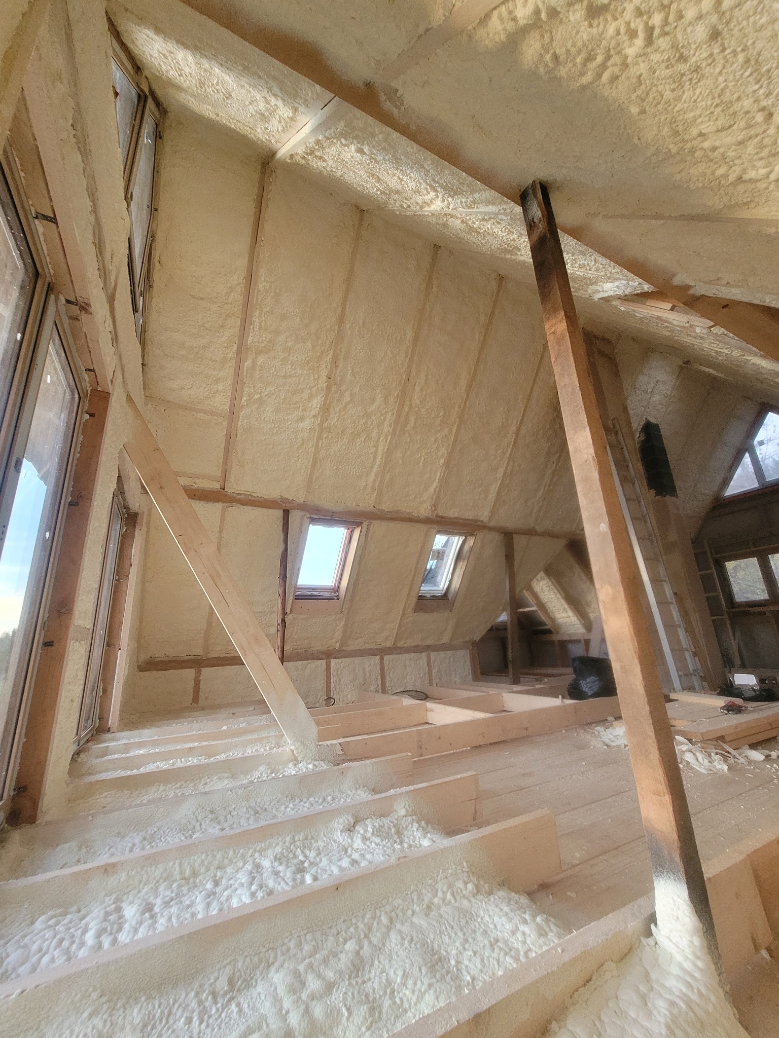 Poddasze po natrysku izolacji. Widoczne drewniane elementy konstrukcyjne i okna. Izolacja pokrywa ściany i sufit, tworząc jednolitą warstwę. Ukończony etap prac.