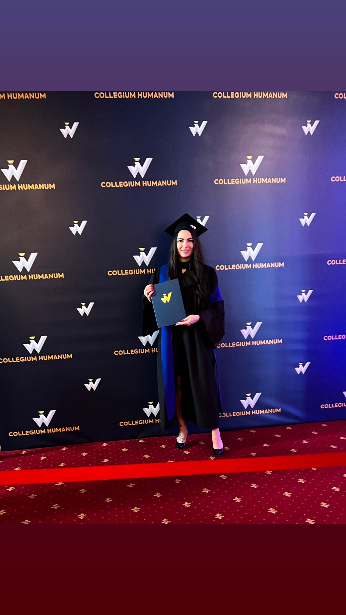 Kobieta w todze i birecie trzyma dyplom na tle ściany z logo Collegium Humanum i czerwonym dywanem.