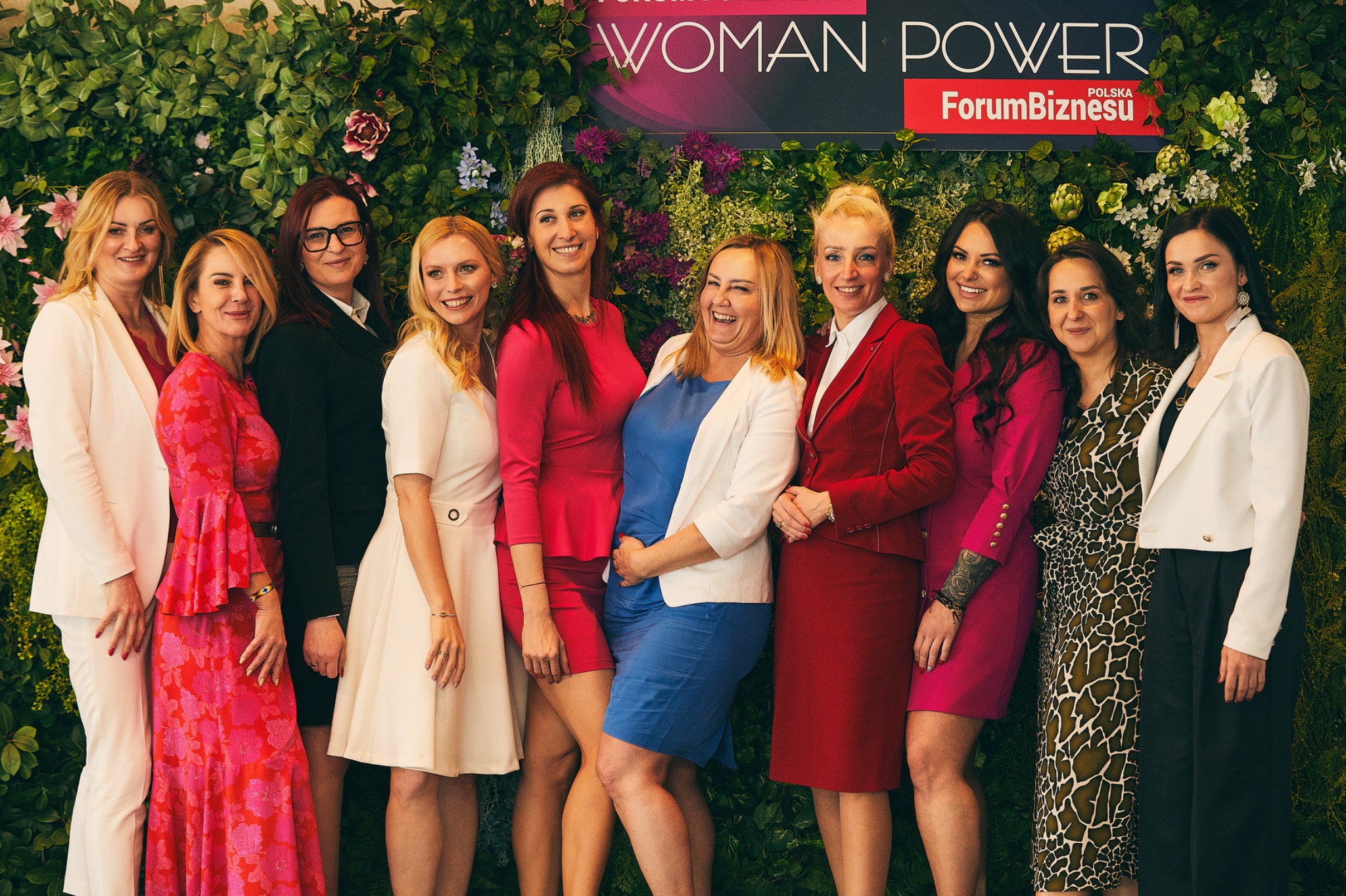 Grupa kobiet w eleganckich strojach pozuje na tle ściany z roślin podczas Forum Biznesu Woman Power Polska.