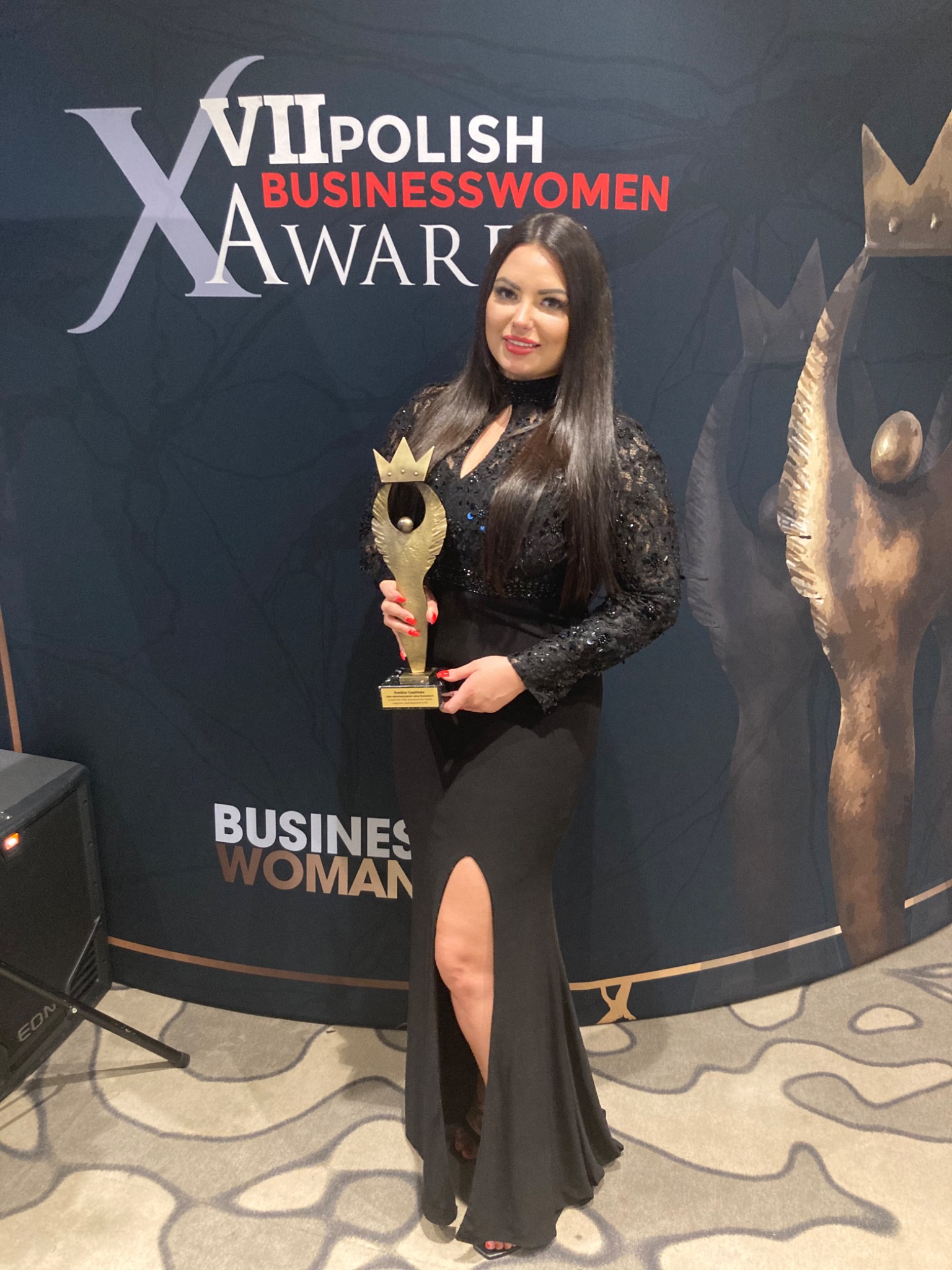 Kobieta w czarnej sukni wieczorowej trzyma złotą statuetkę na tle baneru 'Polish Businesswomen Awards'