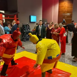 Dwóch mężczyzn w nadmuchiwanych kostiumach sumo walczy na macie podczas wydarzenia w sali kongresowej, w tle widoczni goście i elementy dekoracyjne w stylu japońskim.