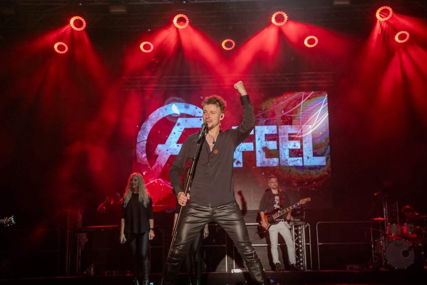 Wokalista z podniesioną ręką śpiewa na scenie w skórzanych spodniach, z tyłu logo zespołu i czerwone reflektory