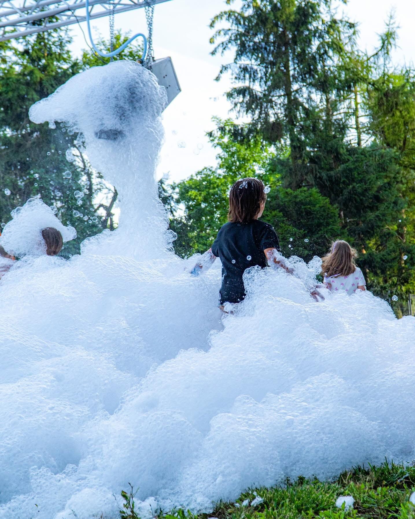 Dzieci bawiące się w pianie generowanej przez maszynę podczas imprezy plenerowej, widok z tyłu na tle zieleni.