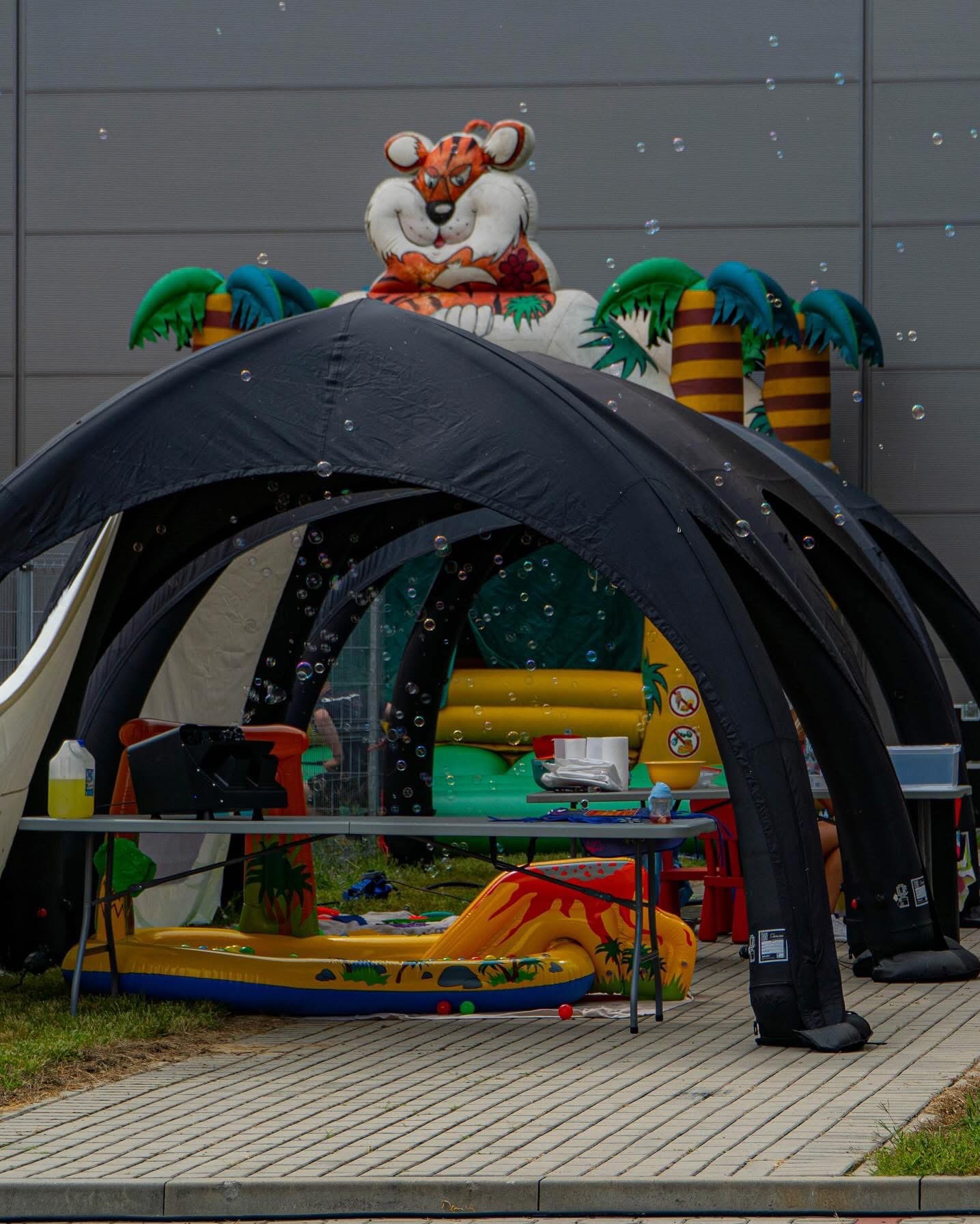 Przygotowania do imprezy plenerowej: dmuchany tygrys i palmy, stół z akcesoriami do puszczania baniek, czarny namiot eventowy, kostka brukowa.