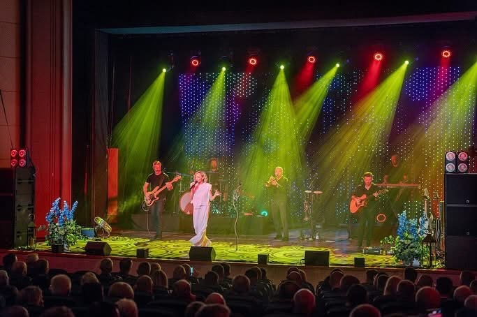 Zespół muzyczny na scenie podczas występu, oświetlony kolorowymi reflektorami, widownia obserwuje koncert.