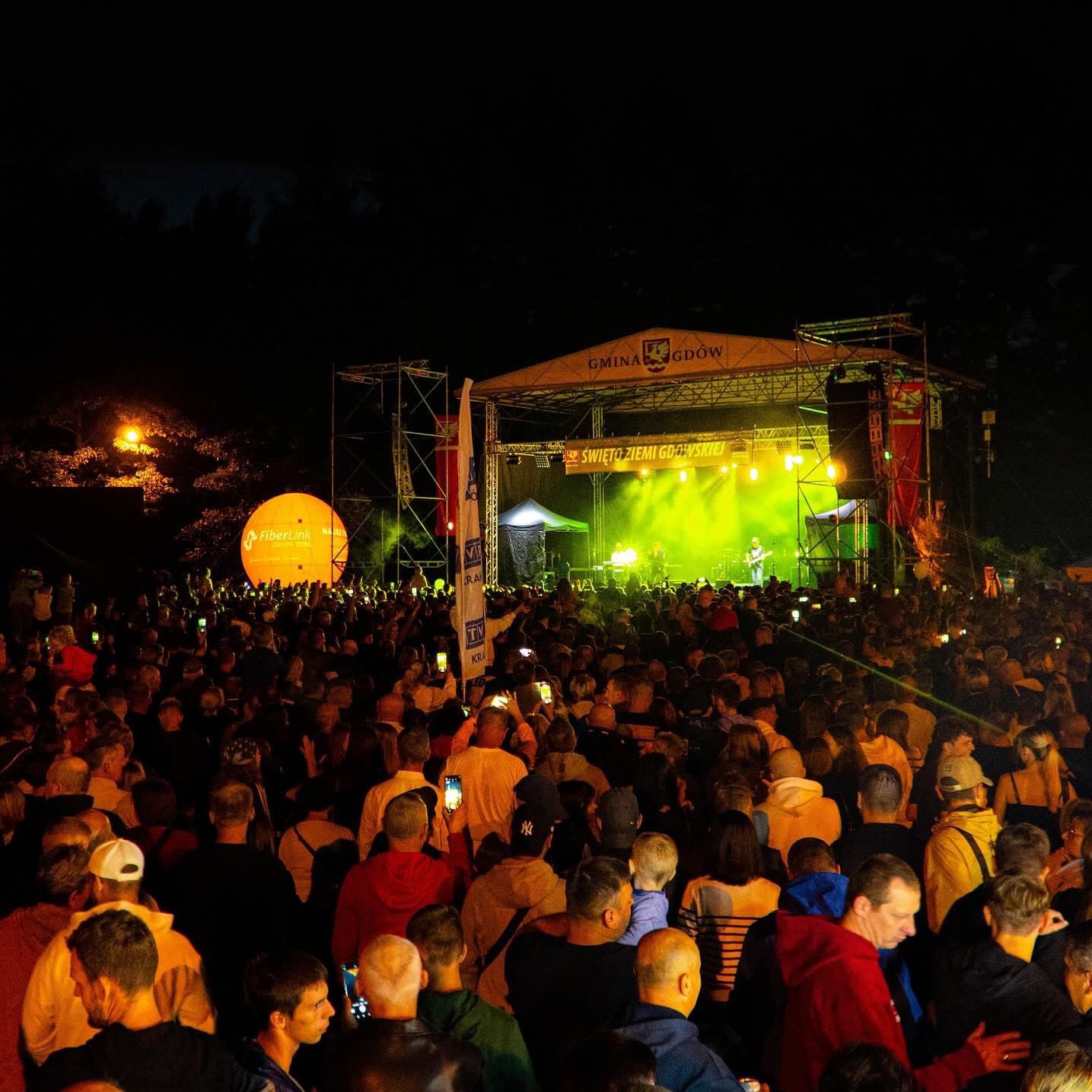 Tłum ludzi obserwujący koncert na scenie oświetlonej zielonym światłem, widoczny baner z napisem 'Święto Ziemi Gdowskiej' oraz duży balon z logo FiberLink.