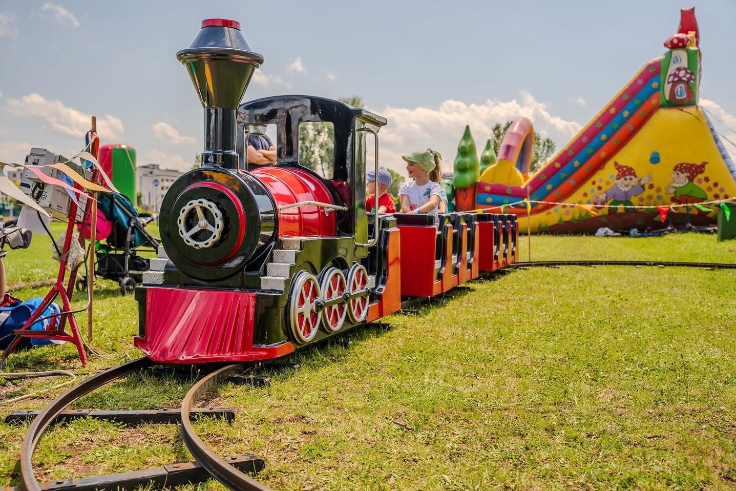 Dzieci jadące w miniaturowej, czerwono-czarnej lokomotywie na torach, z dmuchanym zamkiem w tle, na trawiastym terenie.