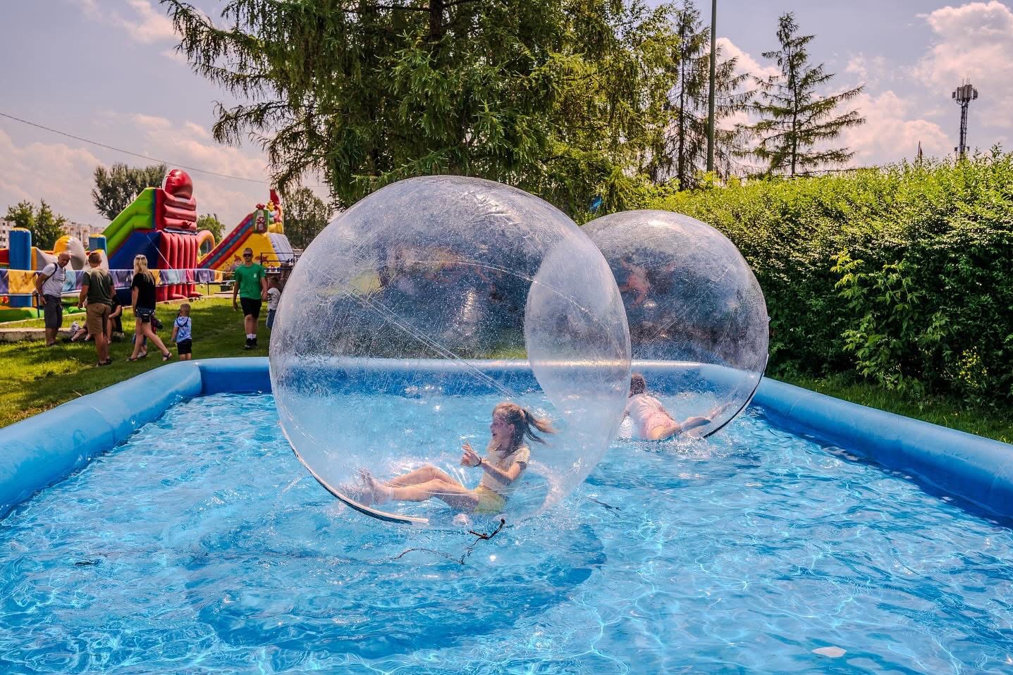 Dwie dziewczynki bawią się wewnątrz przezroczystych, dmuchanych kul w basenie na świeżym powietrzu; w tle dmuchany zamek i inni ludzie.