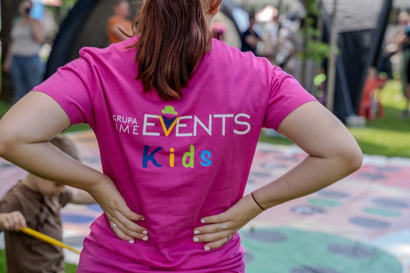 Kobieta w różowej koszulce z napisem 'GRUPA IME EVENTS Kids', widoczna z tyłu, na tle imprezy plenerowej.