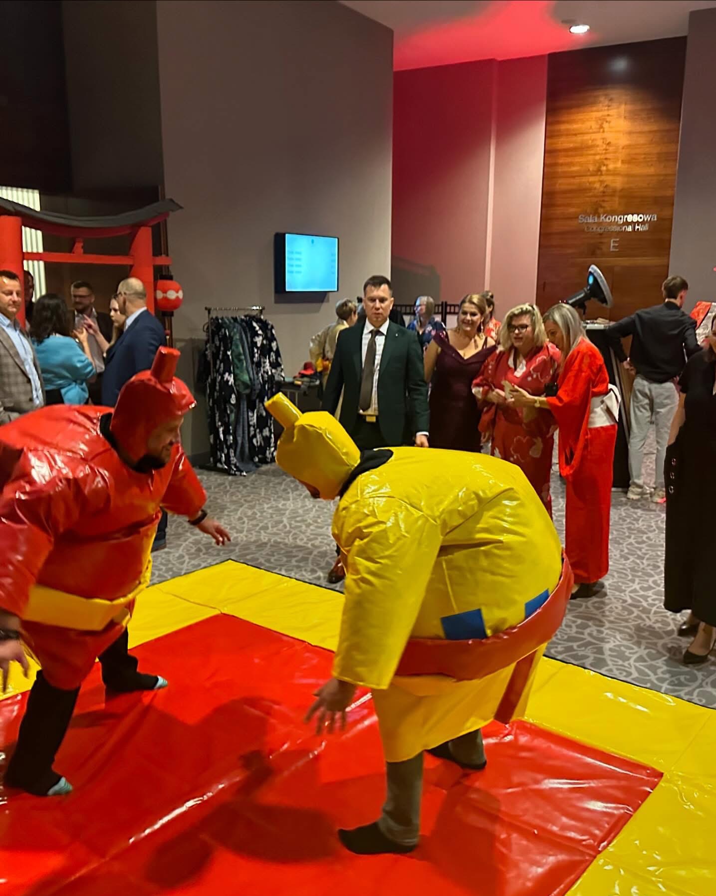 Dwóch mężczyzn w nadmuchiwanych kostiumach sumo walczy na macie podczas wydarzenia w sali kongresowej, w tle widoczni goście i elementy dekoracyjne w stylu japońskim.