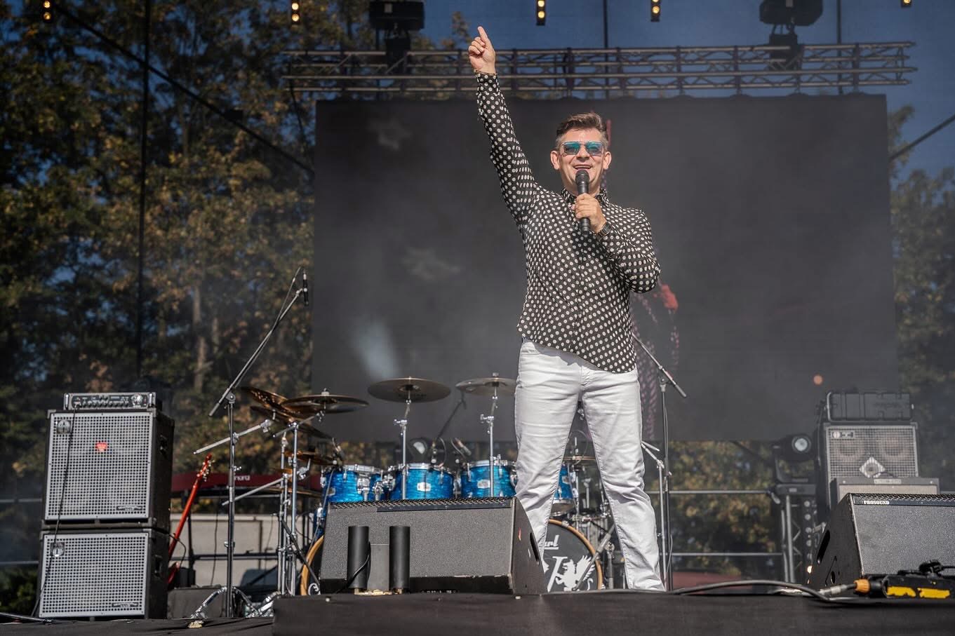 Wokalista w koszuli w grochy z mikrofonem na scenie, w tle perkusja i nagłośnienie, podniesiona ręka w geście powitania.