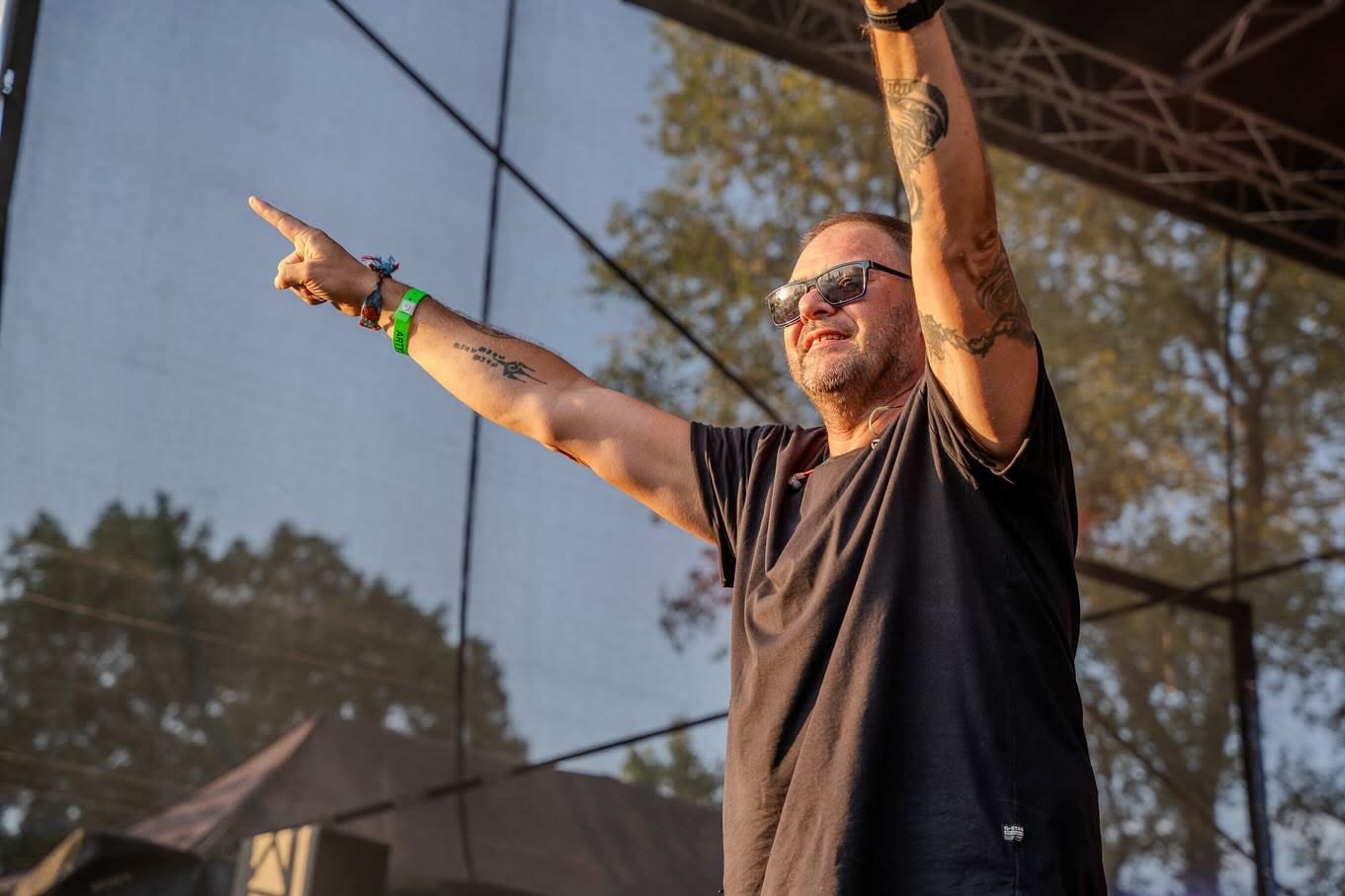 Mężczyzna w czarnej koszulce i okularach przeciwsłonecznych, z tatuażami na ramionach, stoi na scenie i wskazuje palcem w górę, trzymając ręce uniesione, widoczne bransoletki na nadgarstkach.