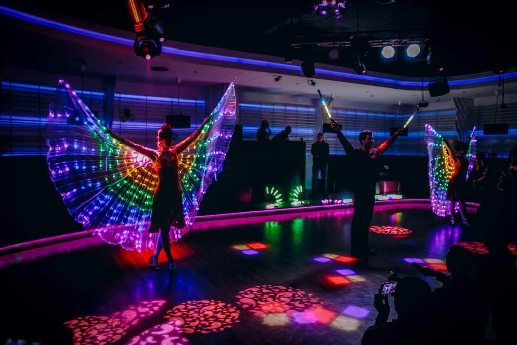 Scena z dwójką performerów w podświetlanych kostiumach, kobieta z tęczowymi skrzydłami i mężczyzna z podświetlanymi pałeczkami, oświetlenie sceniczne w tle, publiczność obserwuje.