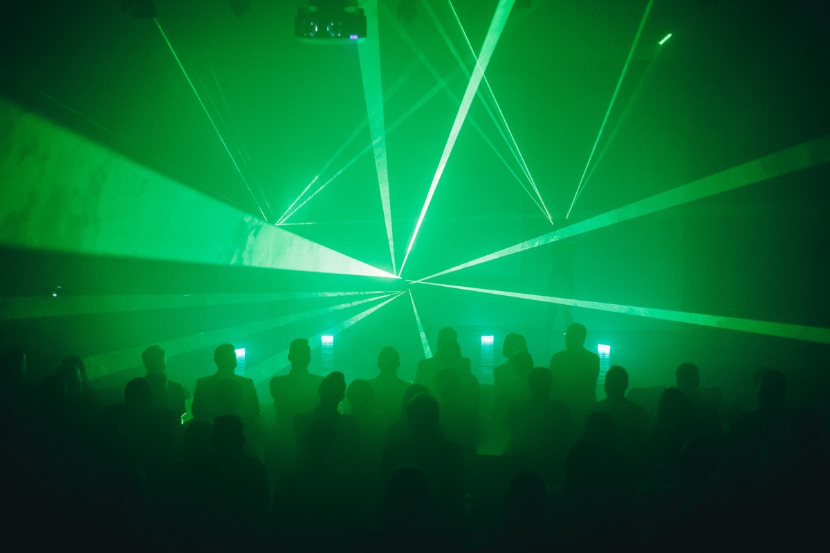 Zielone lasery oświetlają sylwetki publiczności podczas wydarzenia, widoczne projektory na suficie.