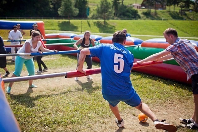 Grupa osób gra w dmuchane piłkarzyki na świeżym powietrzu, widoczny fragment boiska z kolorowymi bandami i uczestnicy trzymający się metalowych rurek.
