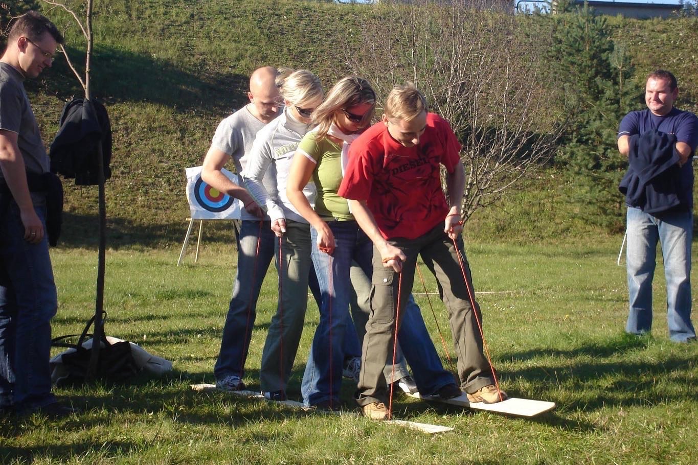 Grupa osób w Krakowie uczestniczy w grze zespołowej na trawie, polegającej na chodzeniu na drewnianych deskach za pomocą sznurków, z tarczą do łucznictwa w tle.