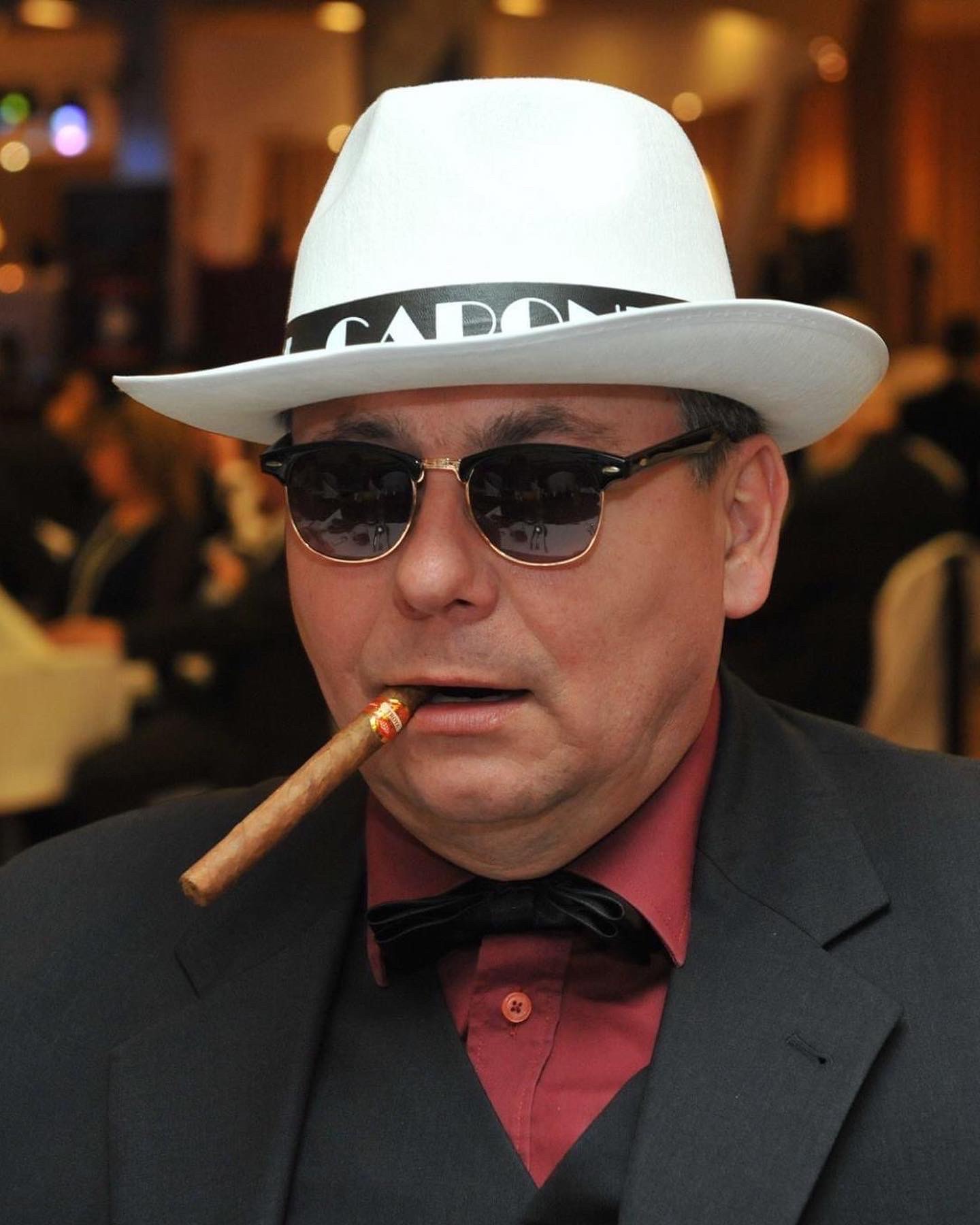 Mężczyzna w białym kapeluszu z czarnym paskiem, okularach przeciwsłonecznych i muszce, palący cygaro w ustach, na tle rozmytego wnętrza.