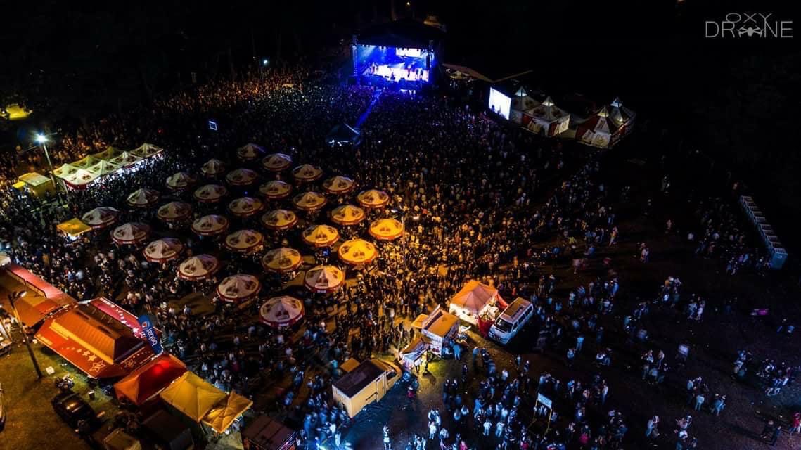 Nocne ujęcie z drona na dużą imprezę plenerową z oświetloną sceną, tłumem ludzi, stoiskami z parasolami i zapleczem technicznym.