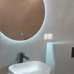 Nowoczesna łazienka z okrągłym lustrem podświetlanym LED, białą umywalką nablatową na drewnopodobnej szafce i czarną baterią, wykończona szarymi płytkami imitującymi marmur.