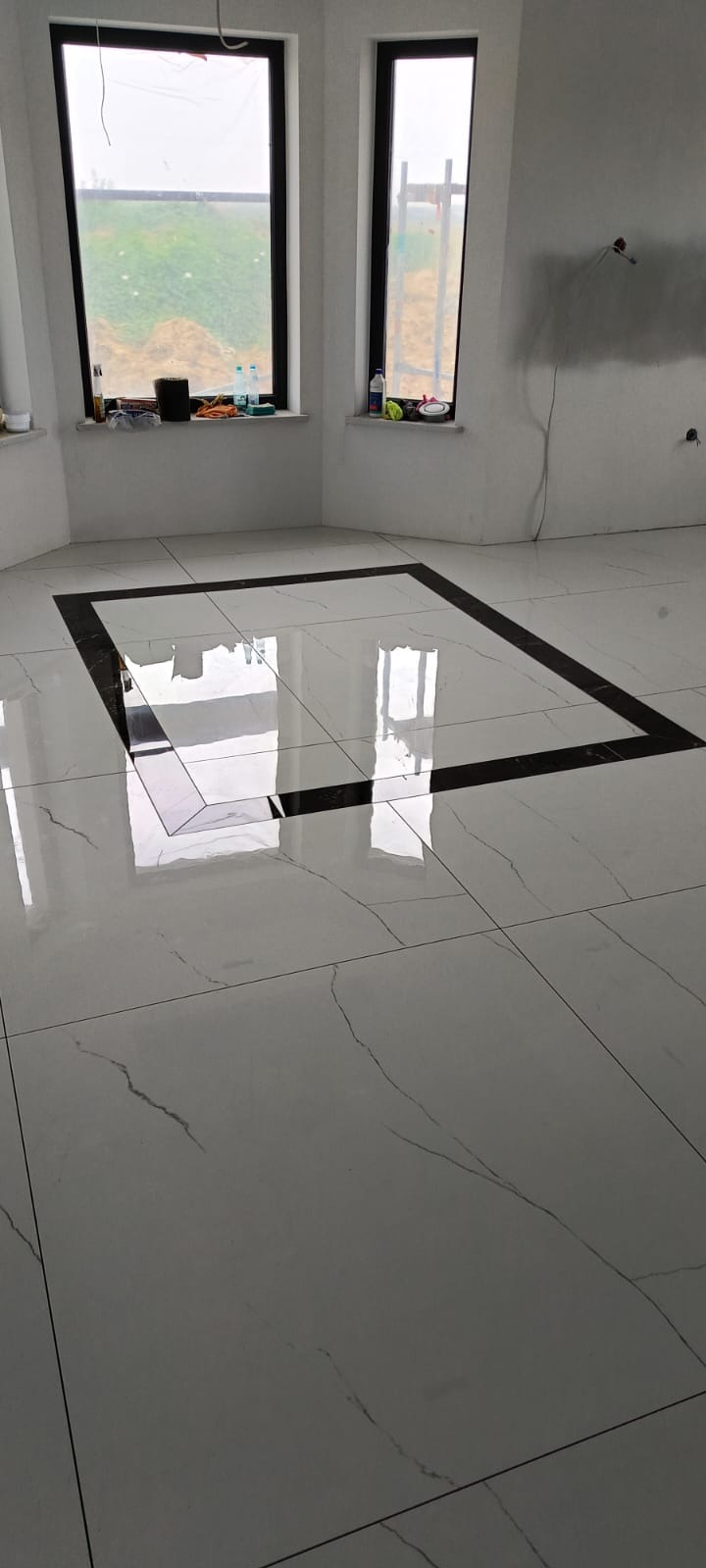 Błyszcząca, biała podłoga z płytek z czarną ramą dekoracyjną w nowym, jasnym pomieszczeniu. Widoczne dwa okna z parapetami.