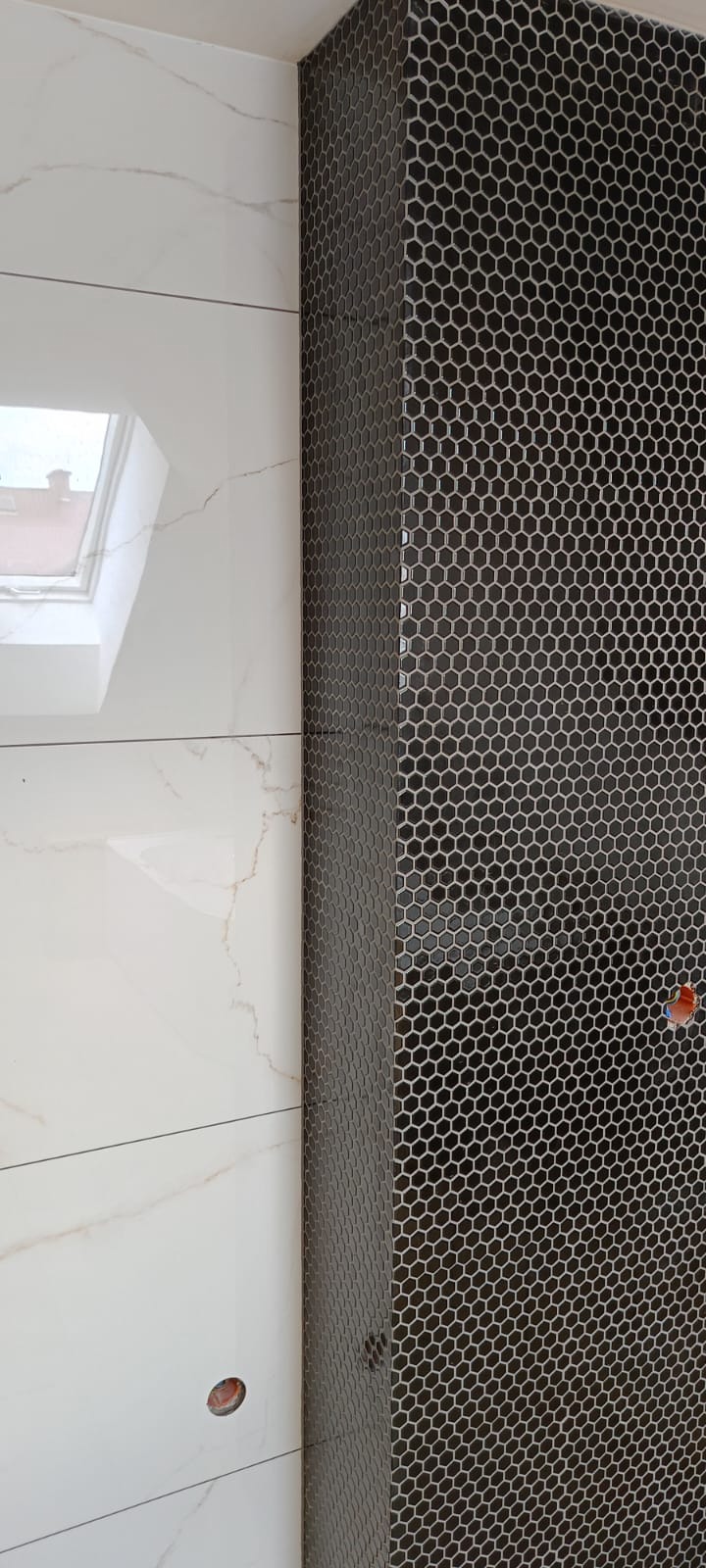 Fragment łazienki z czarną mozaiką heksagonalną na filarze i białymi płytkami imitującymi marmur na ścianie, widoczne okno dachowe w tle.