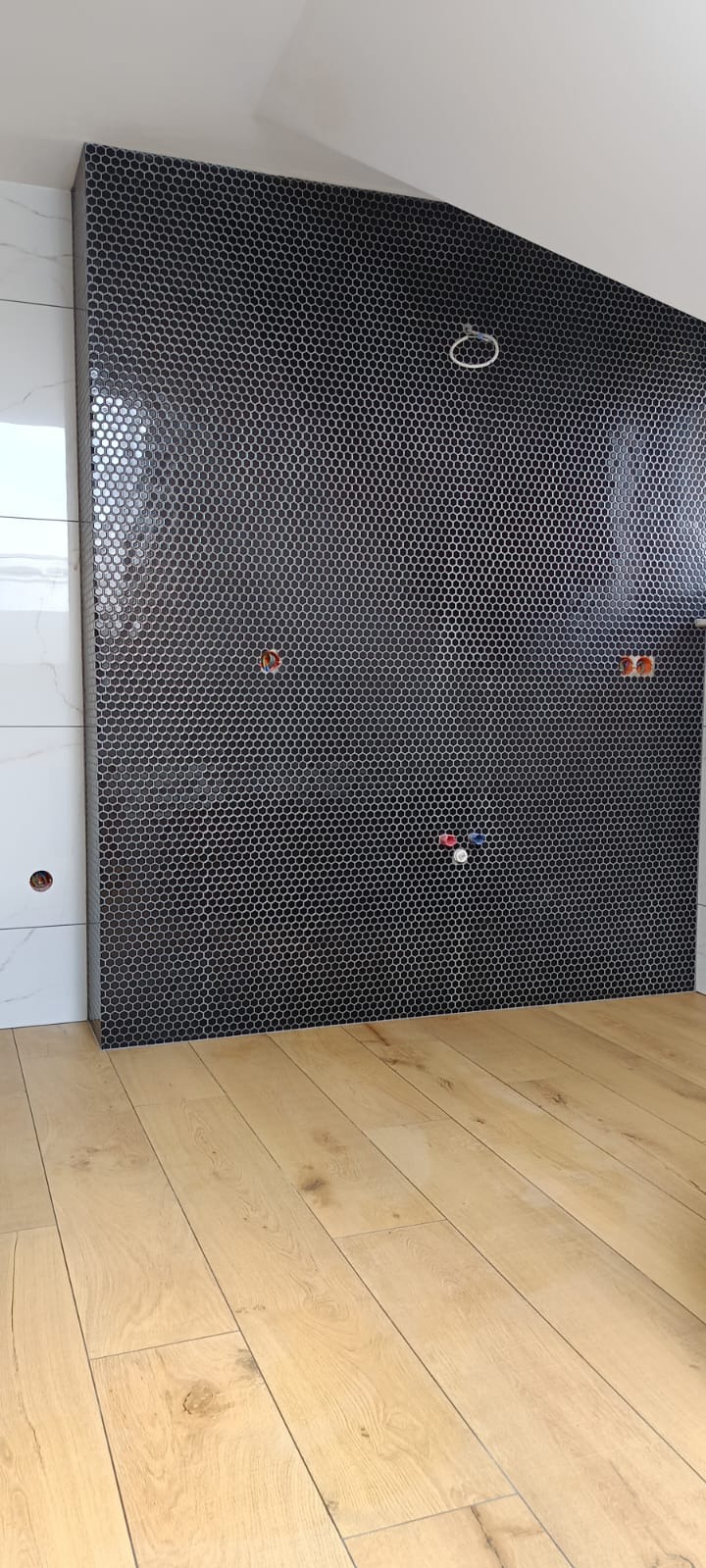 Ściana w łazience wyłożona mozaiką w kształcie czarnych sześciokątów, widoczne przyłącza wodne i elektryczne, podłoga wyłożona panelami drewnopodobnymi.