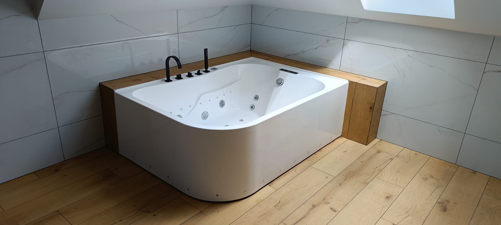 Nowoczesna, biała wanna z hydromasażem w łazience na poddaszu, obudowana drewnem, z czarną armaturą i marmurowymi płytkami na ścianach.