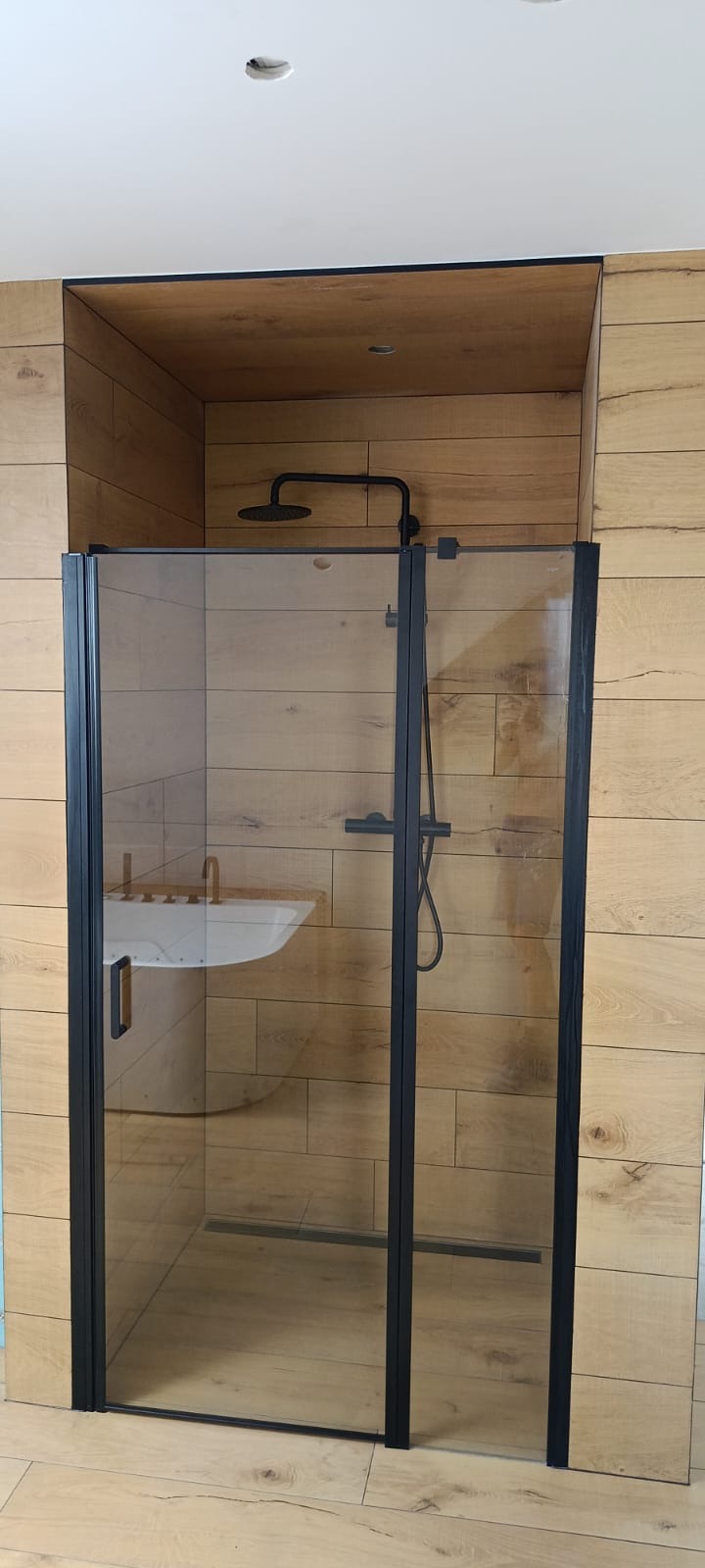 Nowoczesna kabina prysznicowa z czarnymi profilami i deszczownicą, na tle ściany wyłożonej panelami imitującymi drewno, z widoczną umywalką w tle.