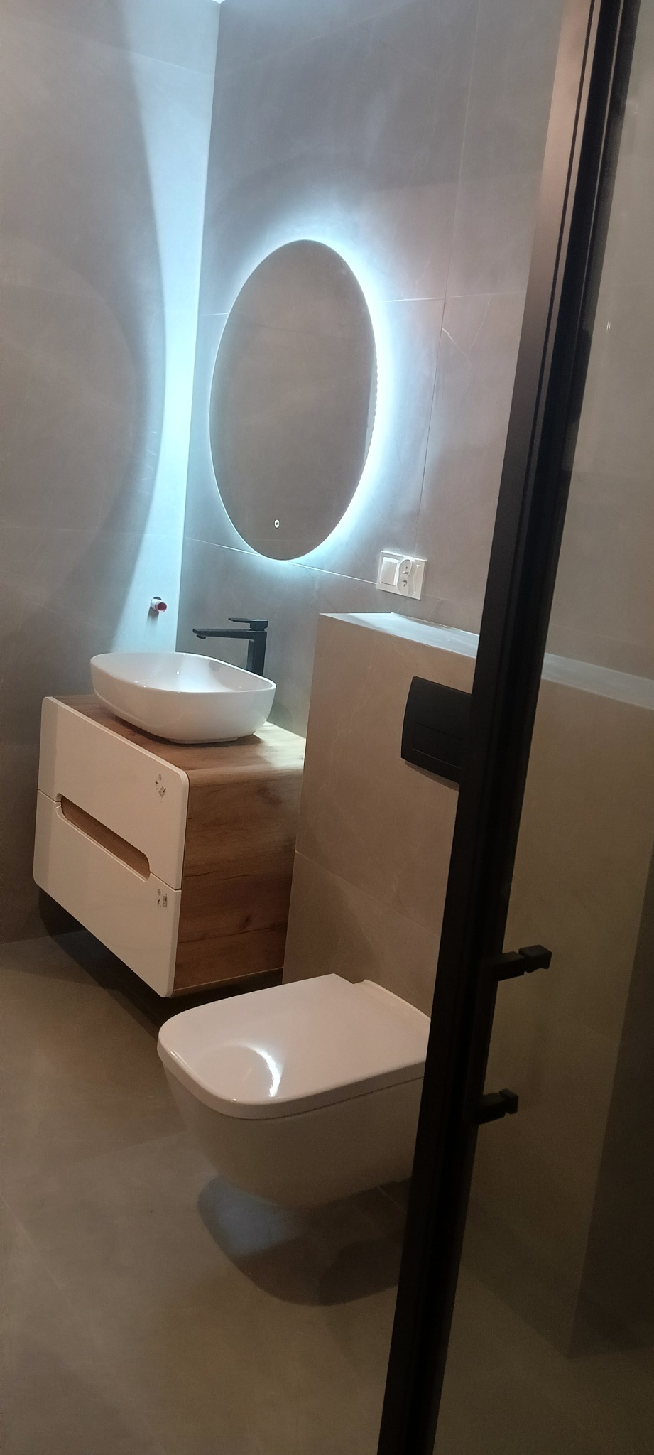 Nowoczesna łazienka z owalnym lustrem z podświetleniem LED, umywalką nablatową na szafce z drewnianym akcentem i wiszącą toaletą, widok zza czarnej ramy kabiny prysznicowej.