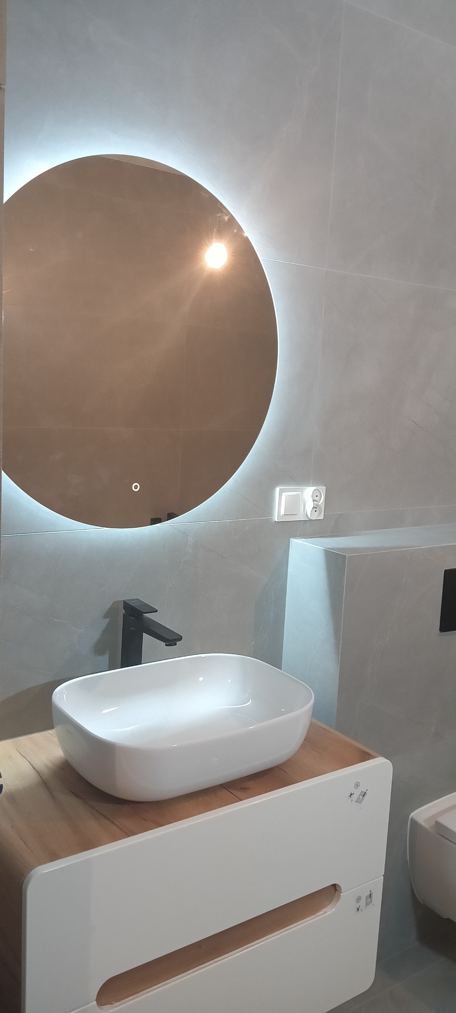 Nowoczesna łazienka z okrągłym lustrem podświetlanym LED, białą umywalką nablatową na drewnopodobnej szafce i czarną baterią, wykończona szarymi płytkami imitującymi marmur.