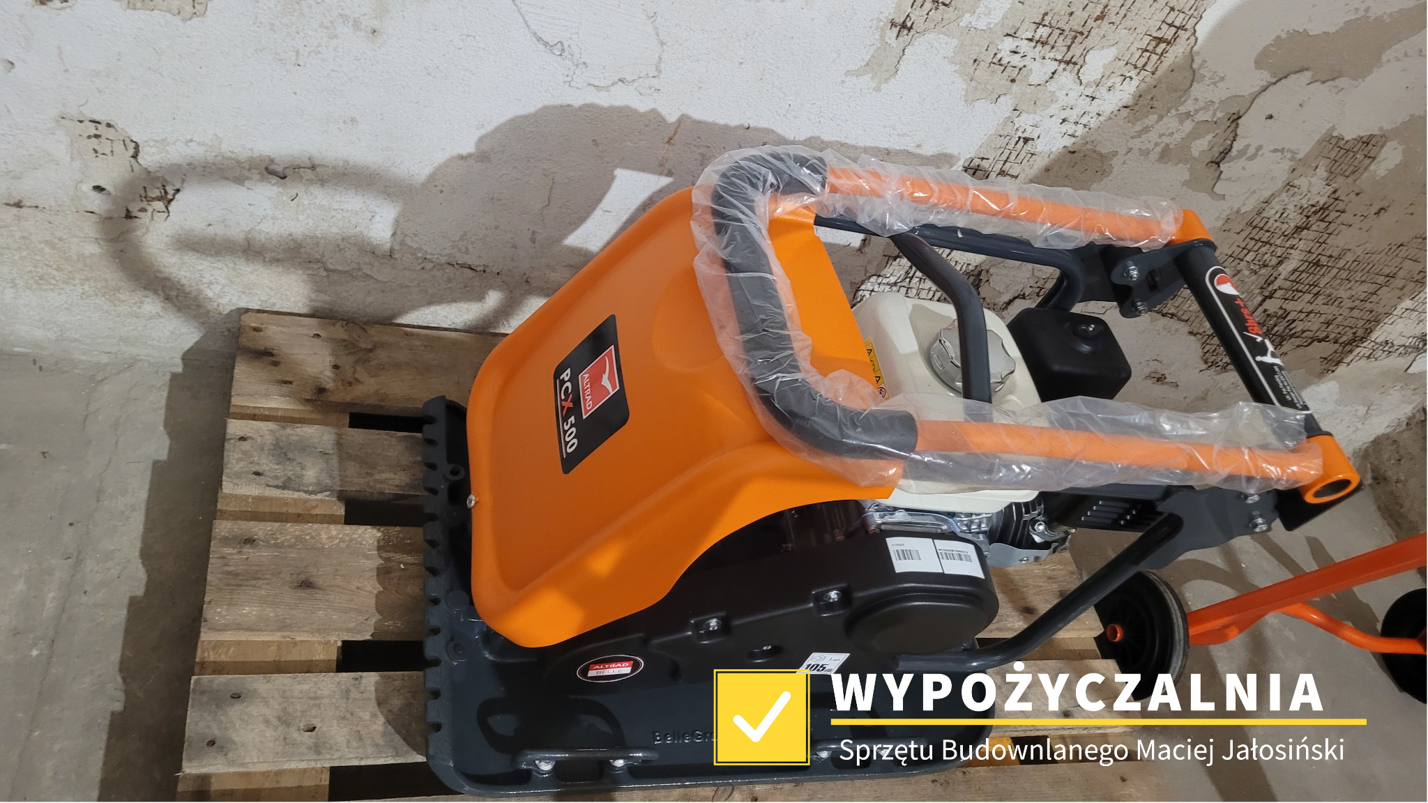 Nowa, pomarańczowo-szara zagęszczarka gruntu PCX 500 Altrad Belle, zapakowana w folię ochronną, stoi na drewnianej palecie na tle ściany z odpadającym tynkiem.