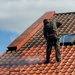 Darfloor - Pracownik w uprzęży, z wysokociśnieniową myjką, czyści czerwoną dachówkę na stromym dachu, z widocznym oknem dachowym i fragmentem drabiny.